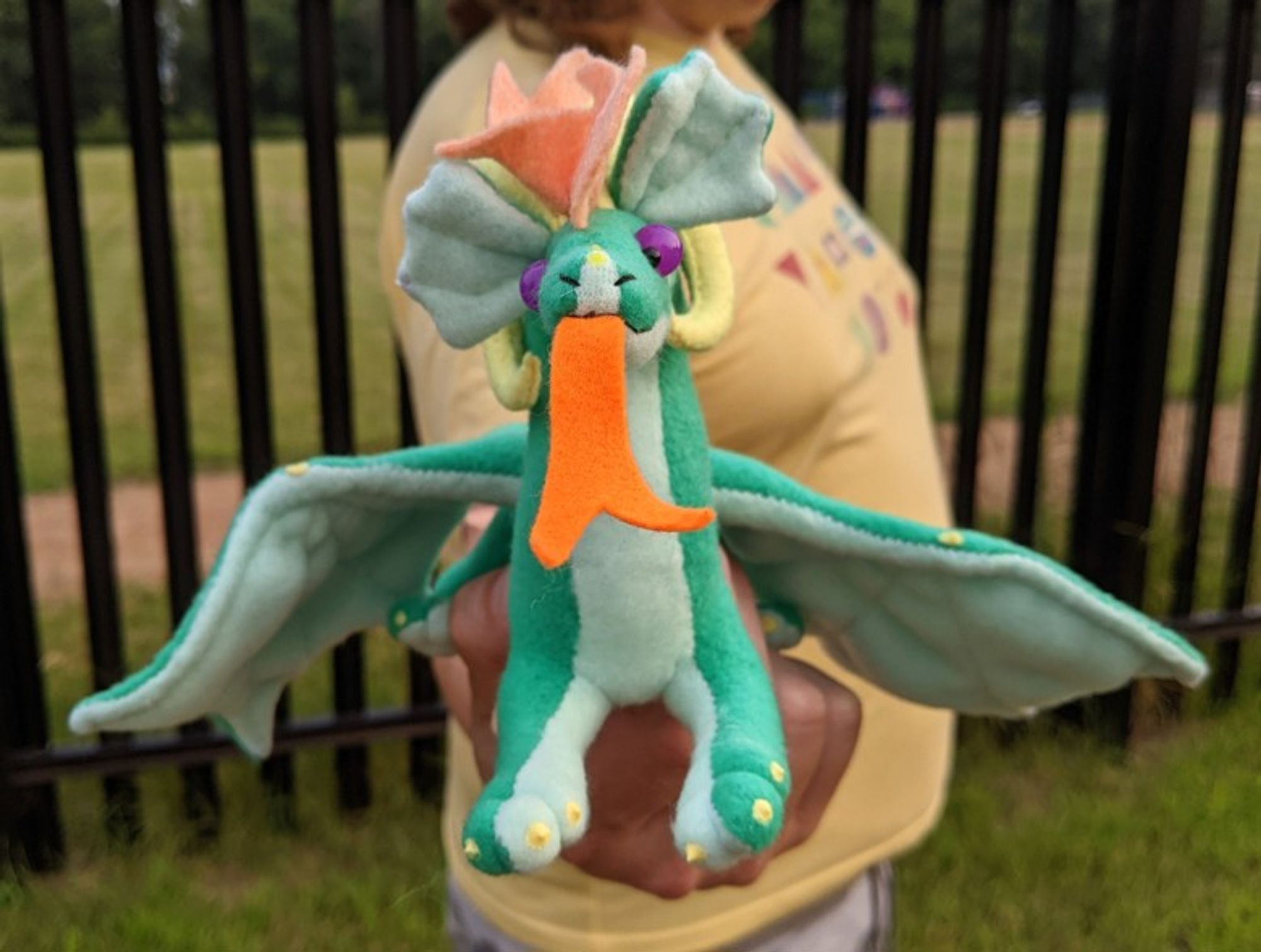 a bottle green dragon plushie with an light aqua underbelly/underwings/ears, a peachy-orange mane/crest and tailtip, yellow ram horns and claws, an orange tongue, and amethyst purple eyes