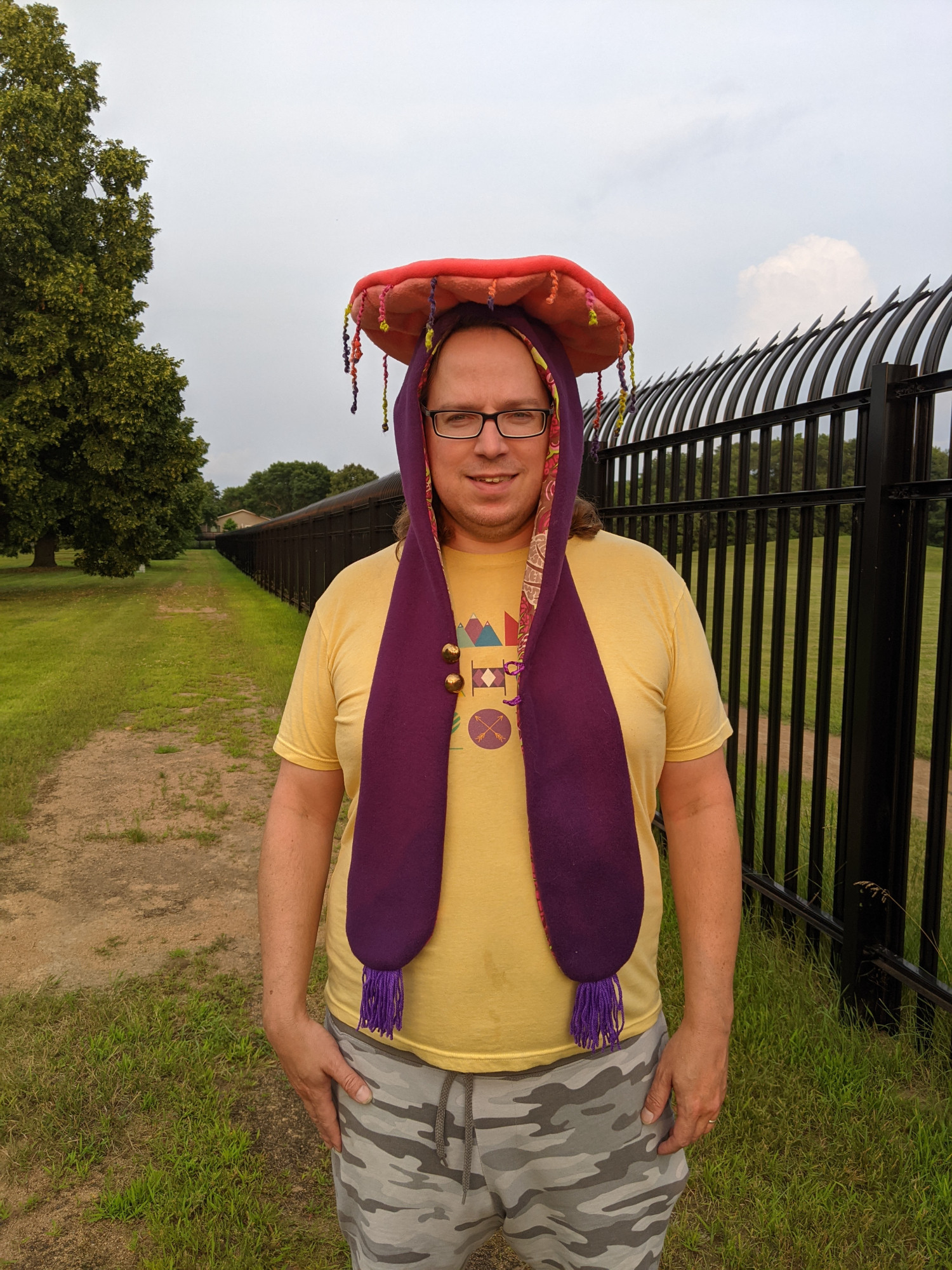 my lovely model Seg wearing the jellyfish hood. it is coral and dark plum-purple fleece with tassels and 2 brass buttons, the tentacles are curly yarn in purple, green, and coral. the liner is pink and green cotton