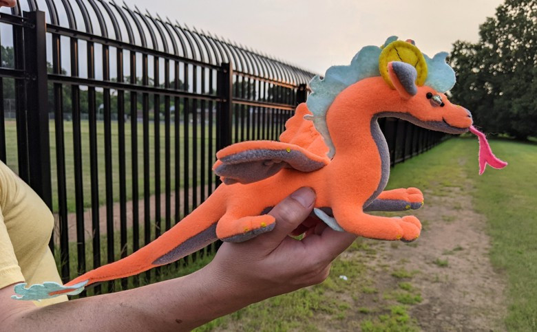 an orange dragon plushie, with a gray underbelly/underwings/inner ears, light blue mane/crest, elbow spines, and tailtip, yellow claws and semicircular horns, a pink tongue, and crystally eyes