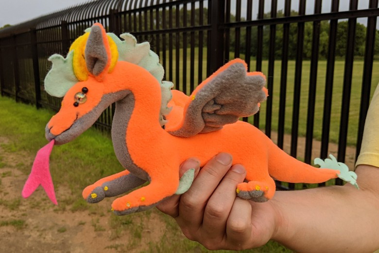 an orange dragon plushie, with a gray underbelly/underwings/inner ears, light blue mane/crest, elbow spines, and tailtip, yellow claws and semicircular horns, a pink tongue, and crystally eyes