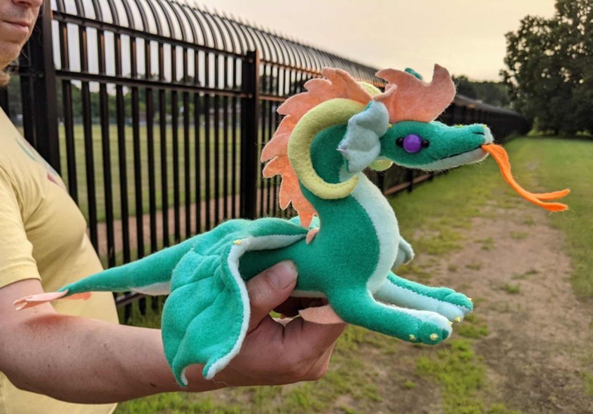 a bottle green dragon plushie with an light aqua underbelly/underwings/ears, a peachy-orange mane/crest and tailtip, yellow ram horns and claws, an orange tongue, and amethyst purple eyes