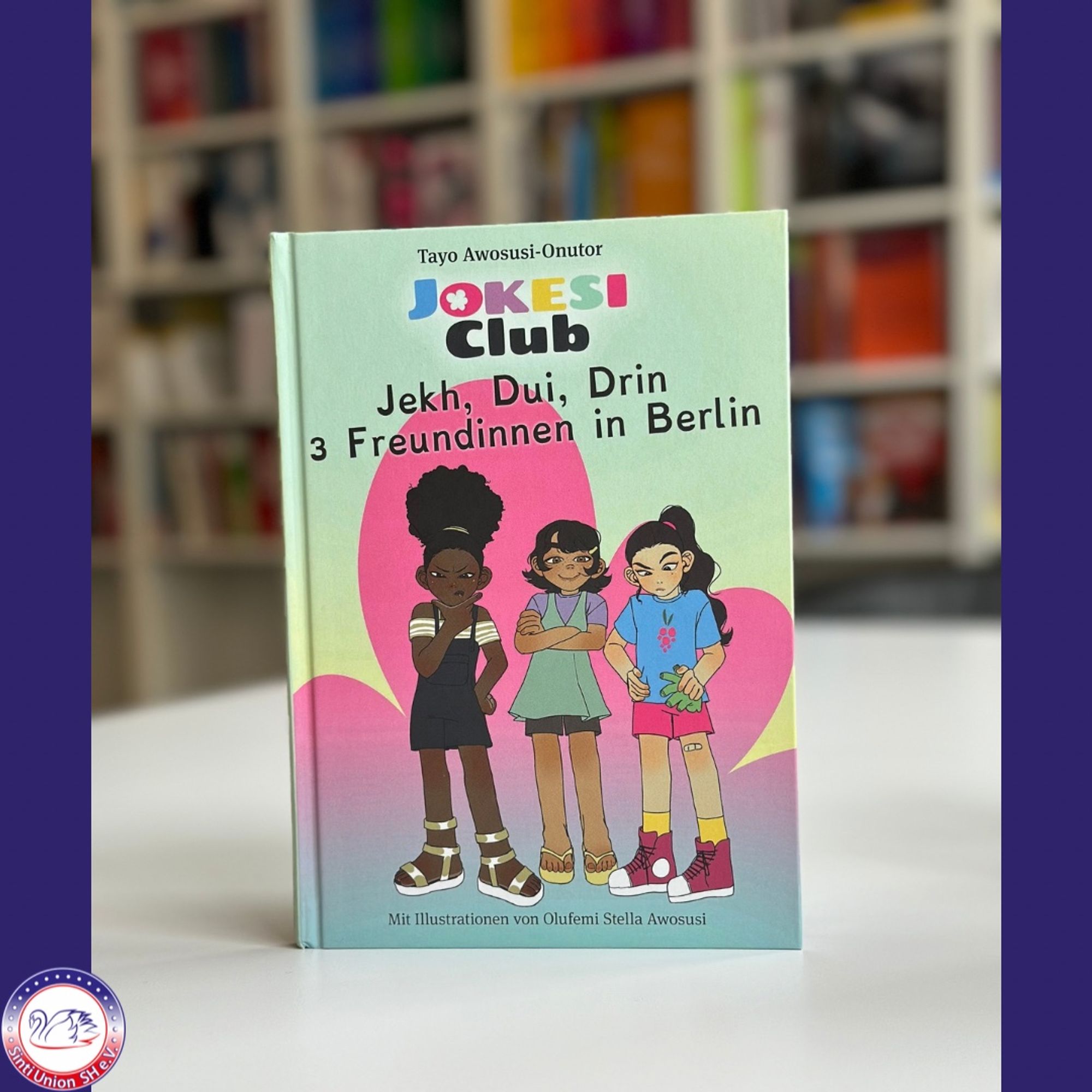 Das Buchcover des Titels „JOKESI Club: Jekh, Dui, Drin – 3 Freundinnen in Berlin“ von Tayo Awosusi-Onutor wird gezeigt. Drei Mädchen, die Hauptcharaktere, stehen selbstbewusst nebeneinander vor einem farbenfrohen Hintergrund. Ihre unterschiedlichen Outfits und Posen lassen auf ihre individuellen Persönlichkeiten schließen. Auch hier wird angegeben, dass die Illustrationen von Olufemi Stella Awosusi gestaltet wurden.