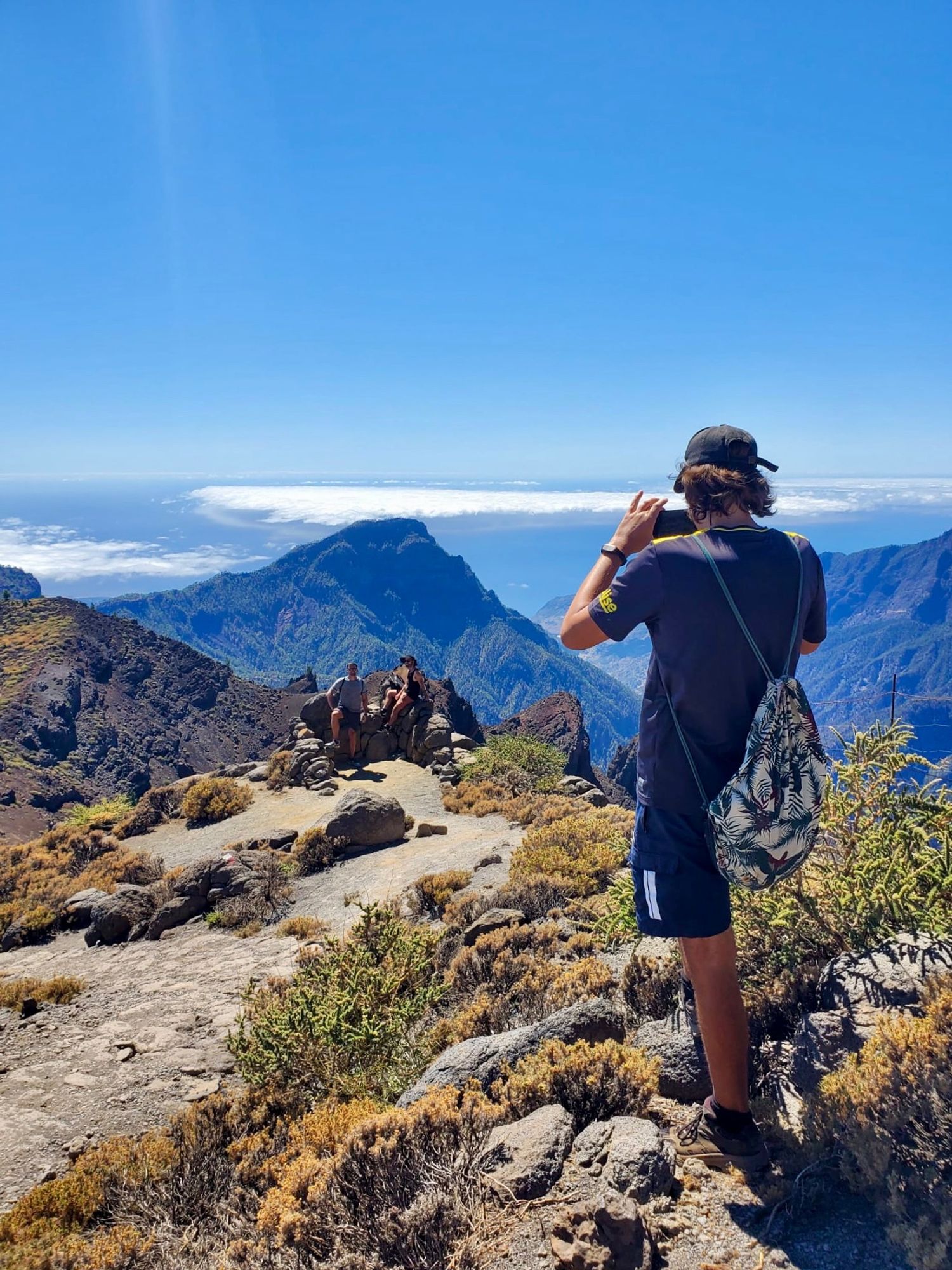 Man taking photo of two fellow hikers, with breathtaking views of La Palma’s volcanoes and super-caldera in the background, with clouds beneath a clear blue sky stretching into space