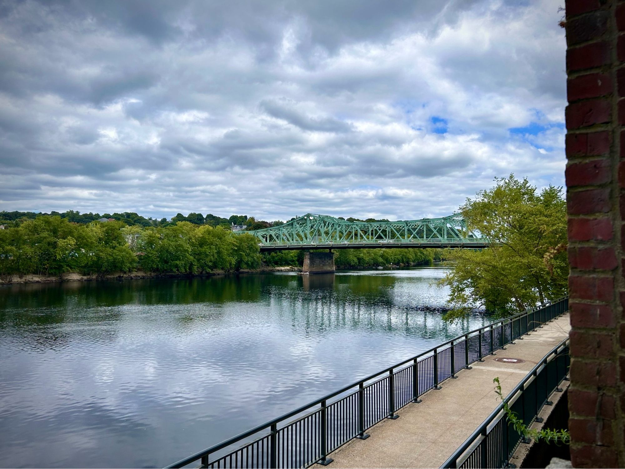 A river with a pale green metal bridge crossing it. Trees on one side and a sidewalk on the other. The sky is mostly cloudy with just a peek of blue