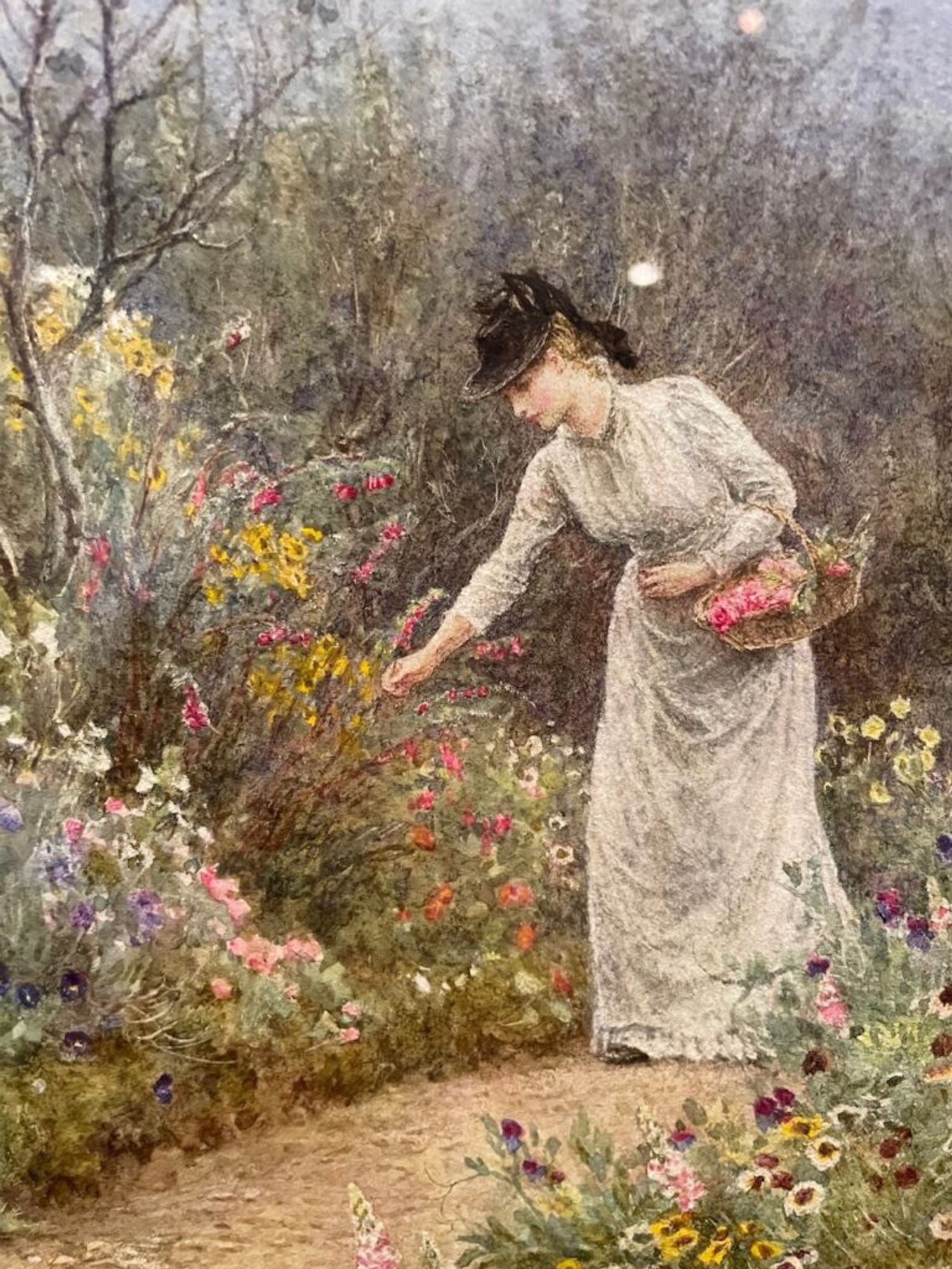 A depiction of a woman in a long white gown and a black hat, with a basket full of cut flowers hooked over her left arm, standing in a path, leaning forward to pick flowers with her right hand.