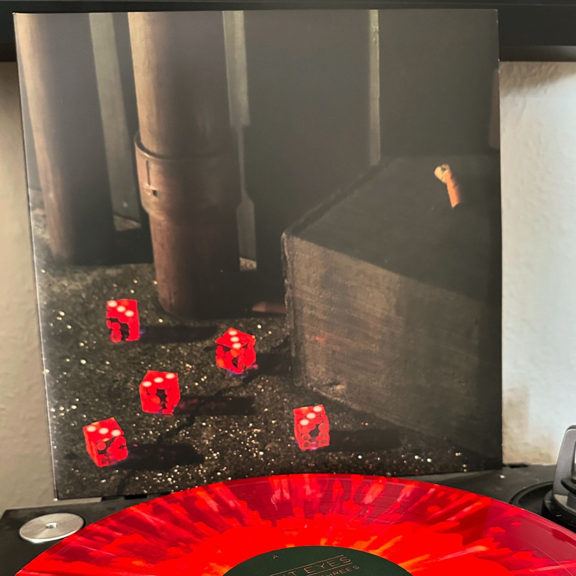 Photo of Bright Eyes’ album “Five Dice, All Threes” on red orange vinyl on a turntable, the album’s cover placed in the background