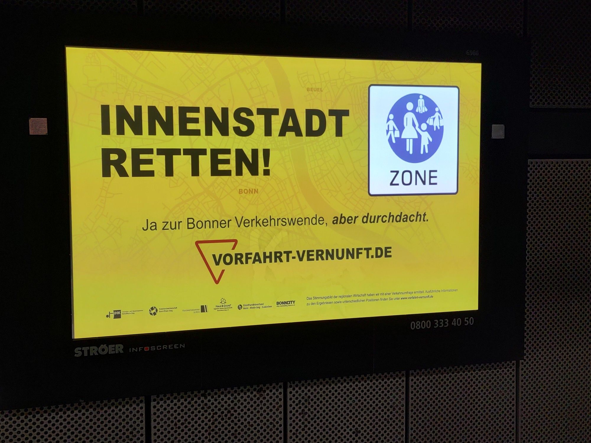 Leuchtreklame in der U-Bahn-Station am Bonner HBF. Auf gelbem Grund: "Innenstadt retten" von "Vorfahrt Vernunft". Dazu ein "Fußgängerzone-Schild" mit mehreren weißen Fußgängern auf blauem Grund