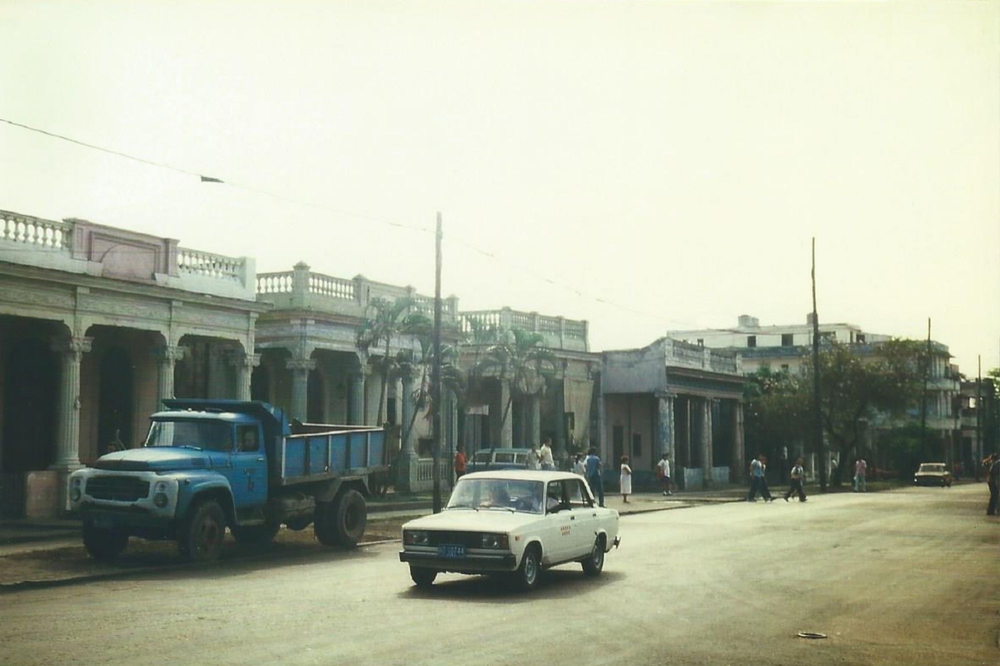 Eine Straße mit Villen im Kolonialstil. Ein heller PKW auf einsamer Straße, halb auf dem Gehweg ein türkisblauer Lastwagen mit Ladefläche. Im Hintergrund laufen einzelne Menschen. Diesiger Himmel.