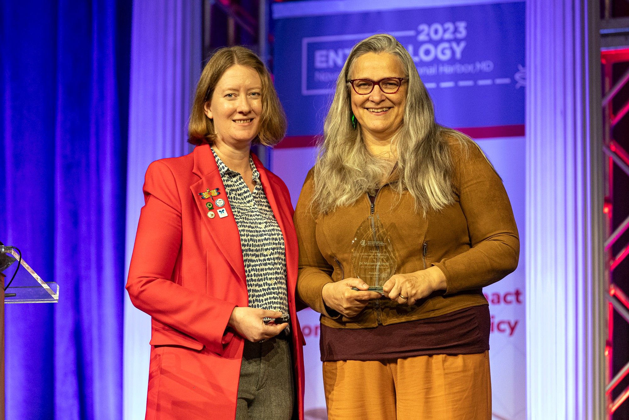 Incoming ESA president Jennifer Henke poses with outgoing president Marianne Alleyne on the main stage at Entomology 2023, after presenting Alleyne with a small crystal statue with the ESA logo, which Alleyne holds.