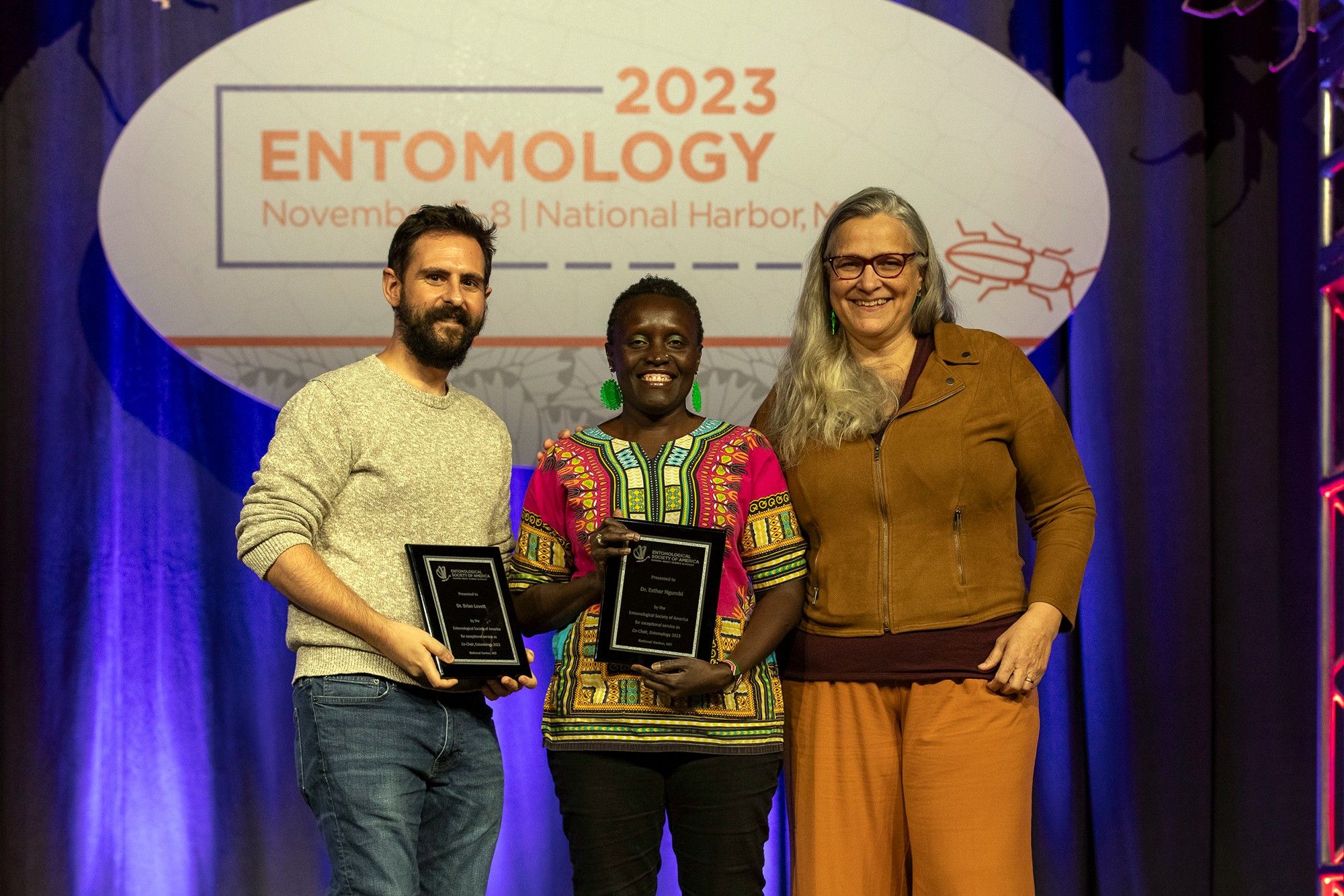 Entomology 2023 program co-chairs Brian Lovett and Esther Ngumbi pose on stage with ESA president Marianne Alleyne. Lovett and Ngumbi are both holding black plaques with silver writing.