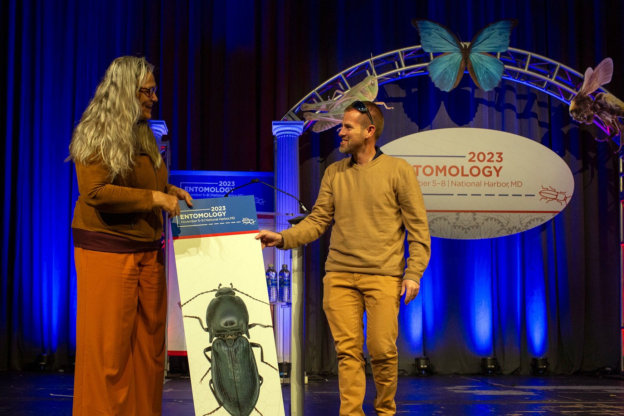 ESA board member Julien Saguez hands outgoing president Marianne Alleyne a tall sign that had adorned the front of the podium on the main stage at Entomology 2023. The sign shows the conference logo in white on blue on the top, with a large image of a black beetle oriented vertically on the sign below.