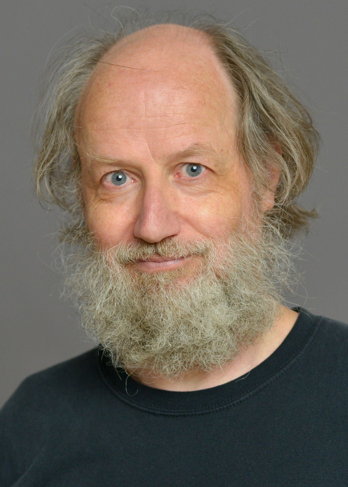 A man with gray hair and a full, graying beard is shown against a plain background. He is wearing a black shirt and has blue eyes.