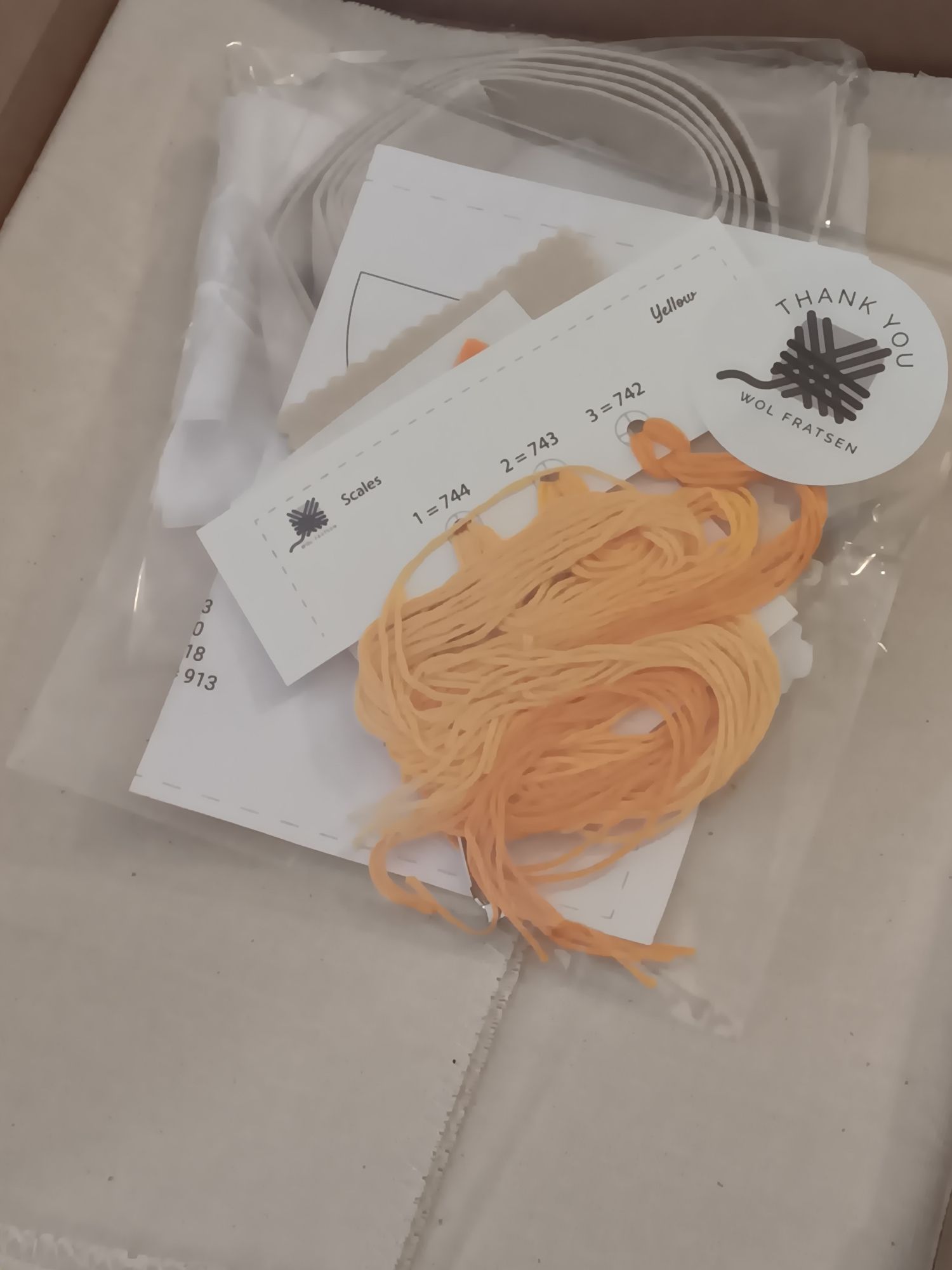 Some orange-yellow embroidery floss and card stock in cellophane and tote bag canvas in a cardboard mailer. There is a THANK YOU WOL FRATSEN stocker on the cellophane.