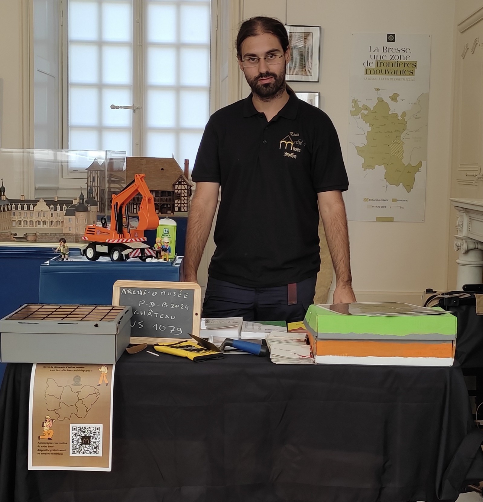 Stand avec des livres, des outils (truelle, outils de dentiste), décor de chantier en figurines Playmobil dans une salle avec une maquette de château