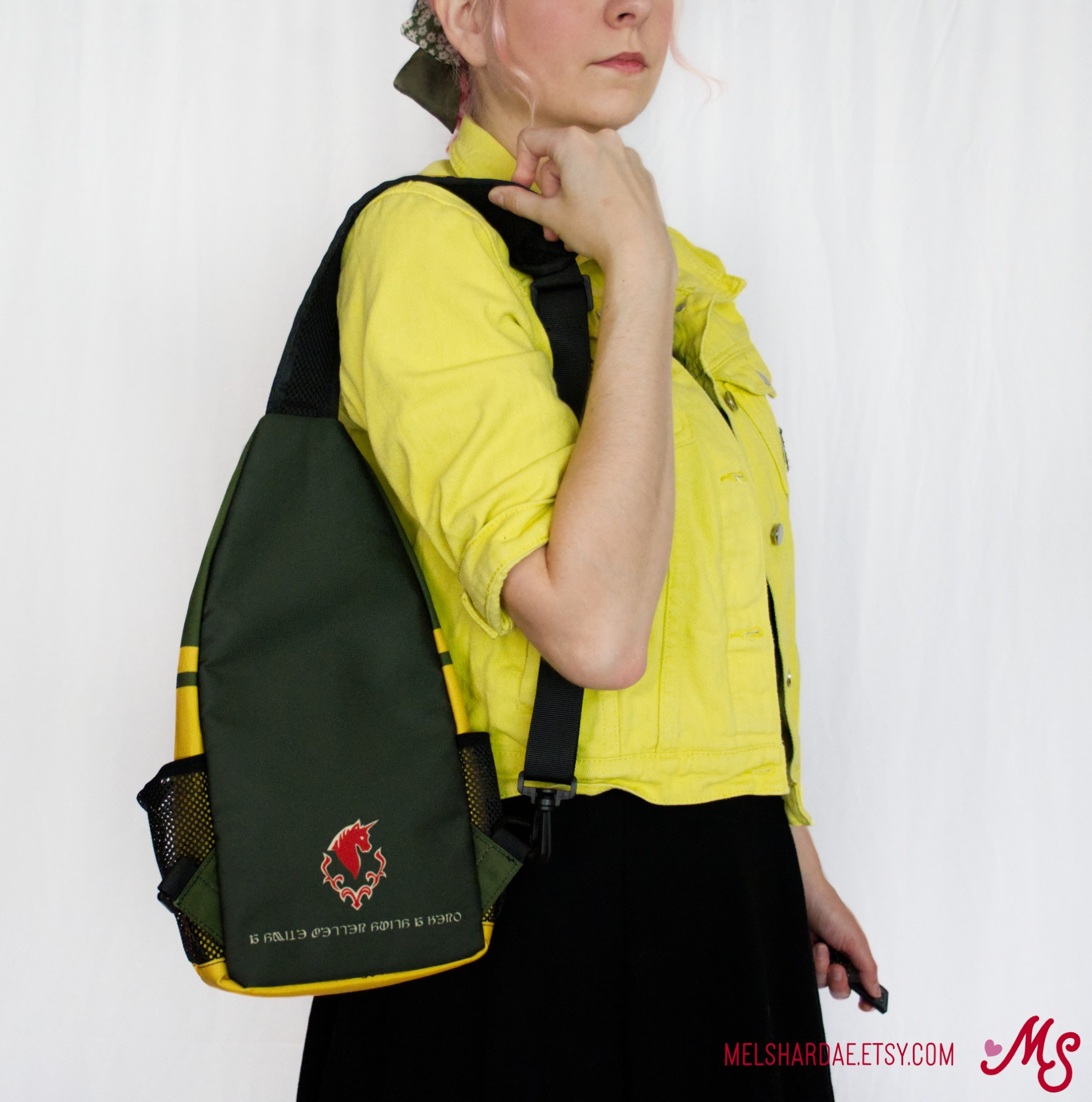 Photo of me modeling my Haurchefant inspired sling bag. It is olive green and grey with yellow accents. The back has a small house Fortemps emblem with the text "a smile better suits a hero" in eorzean 