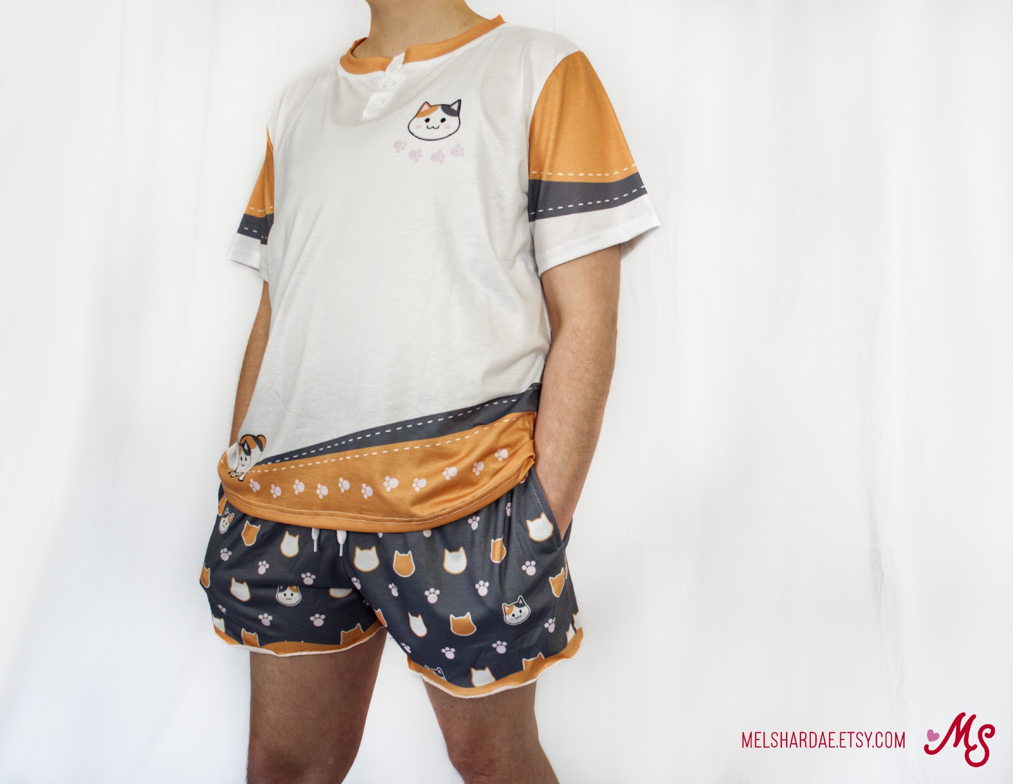 Photo of a model wearing the Fat Cat pajama set. The shirt is white with orange-brown and grey accents and a fat cat on the chest and lower left, and the shorts are dark grey with a repeating fat cat head pattern.