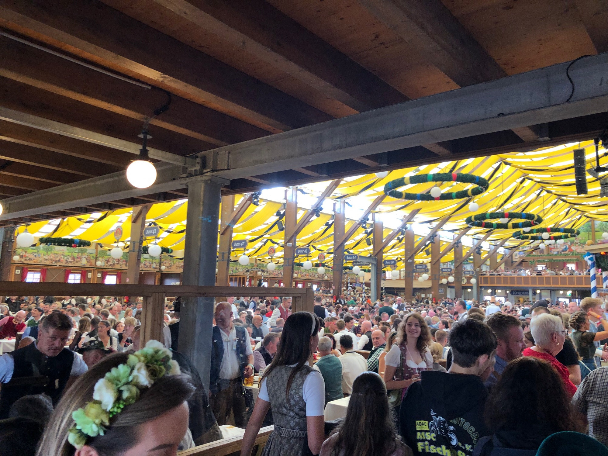 A beer hall with yellow banners on the roof, and circular wreaths hanging below them. Thousands gather to drink beer a d eat the Wurst food you can think of