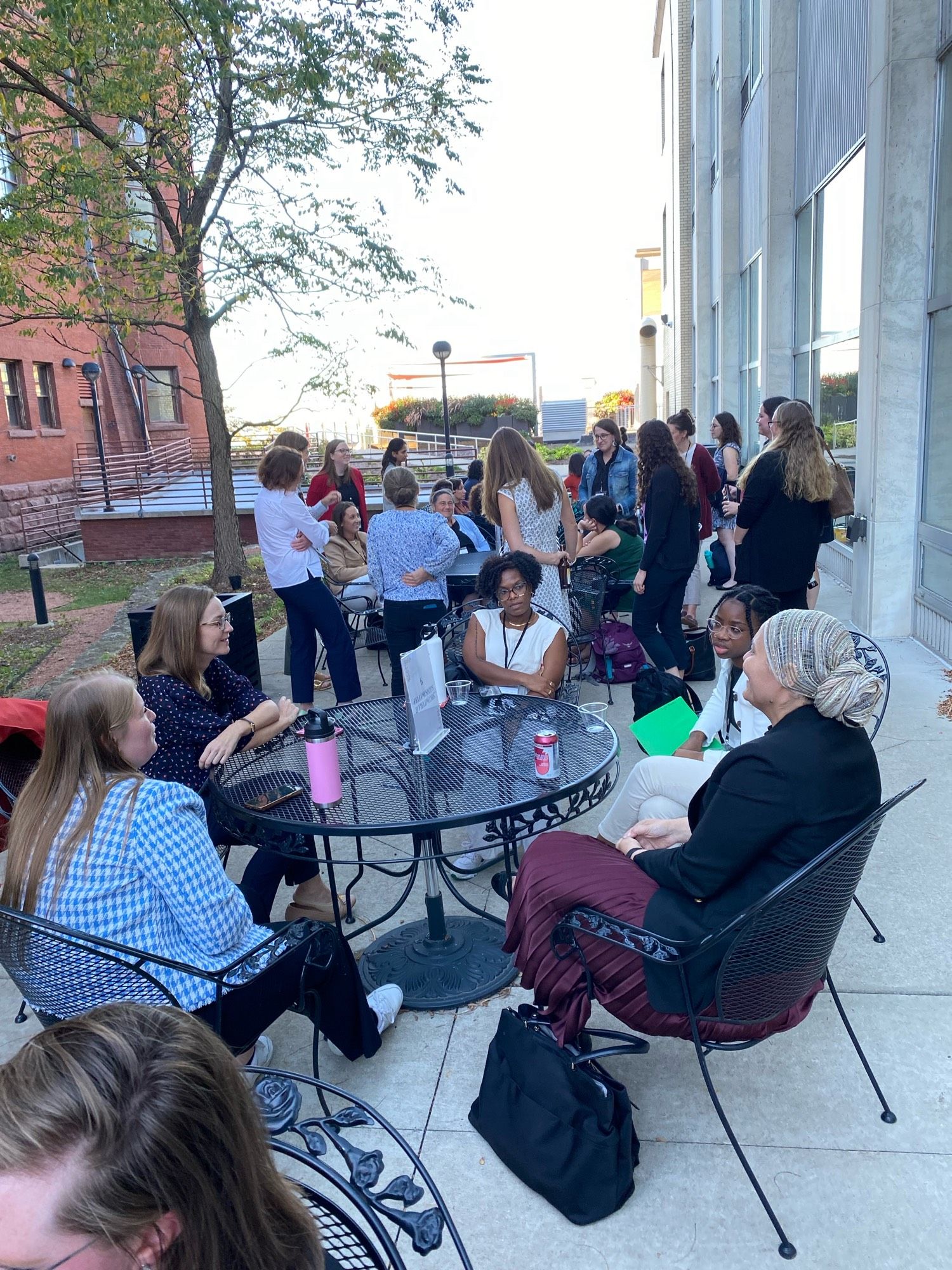 Women stand and sit at an outdoor patio with a small tree and a brick building.