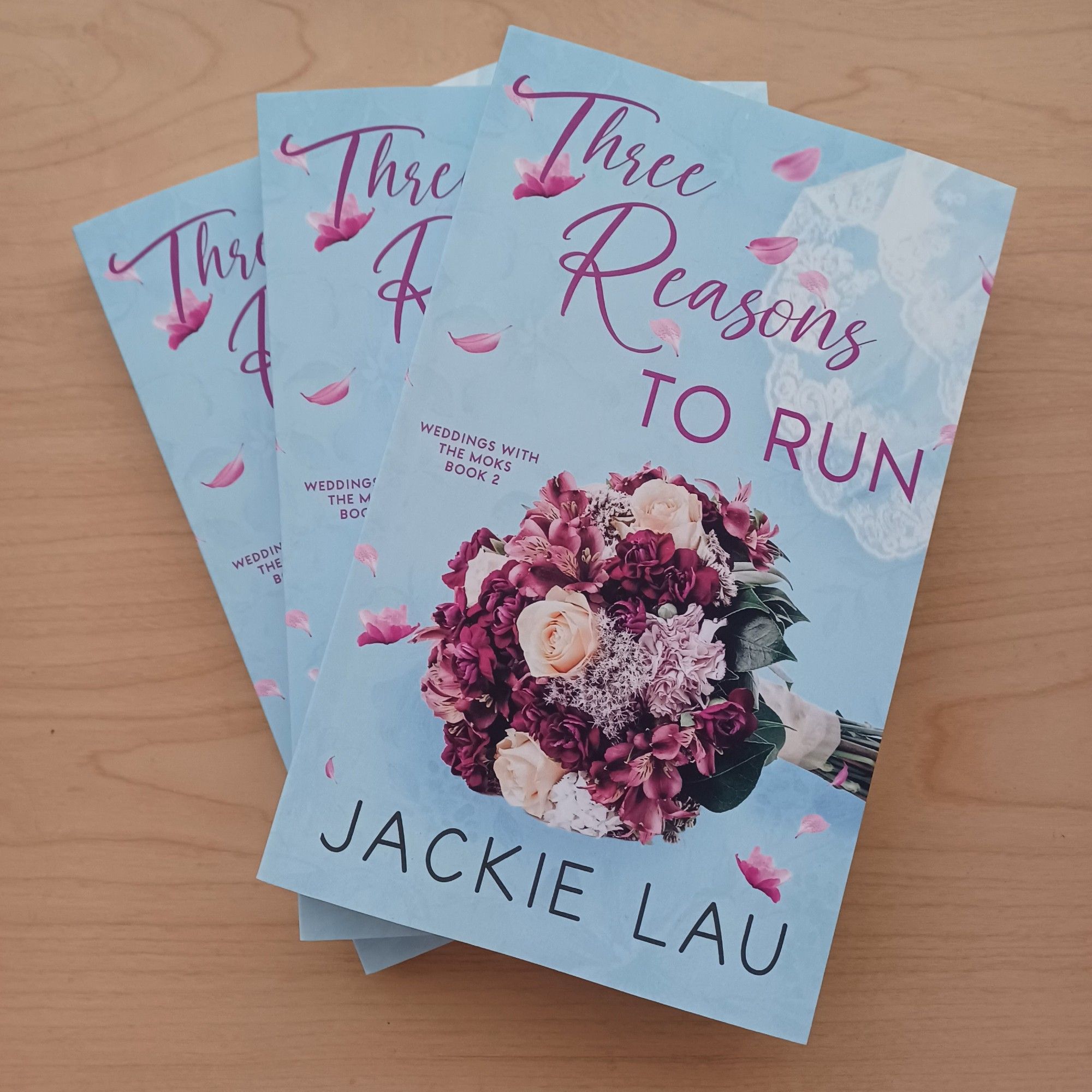 Three fanned paperbacks of Three Reasons to Run by Jackie Lau. Blue cover with bouquet in center.