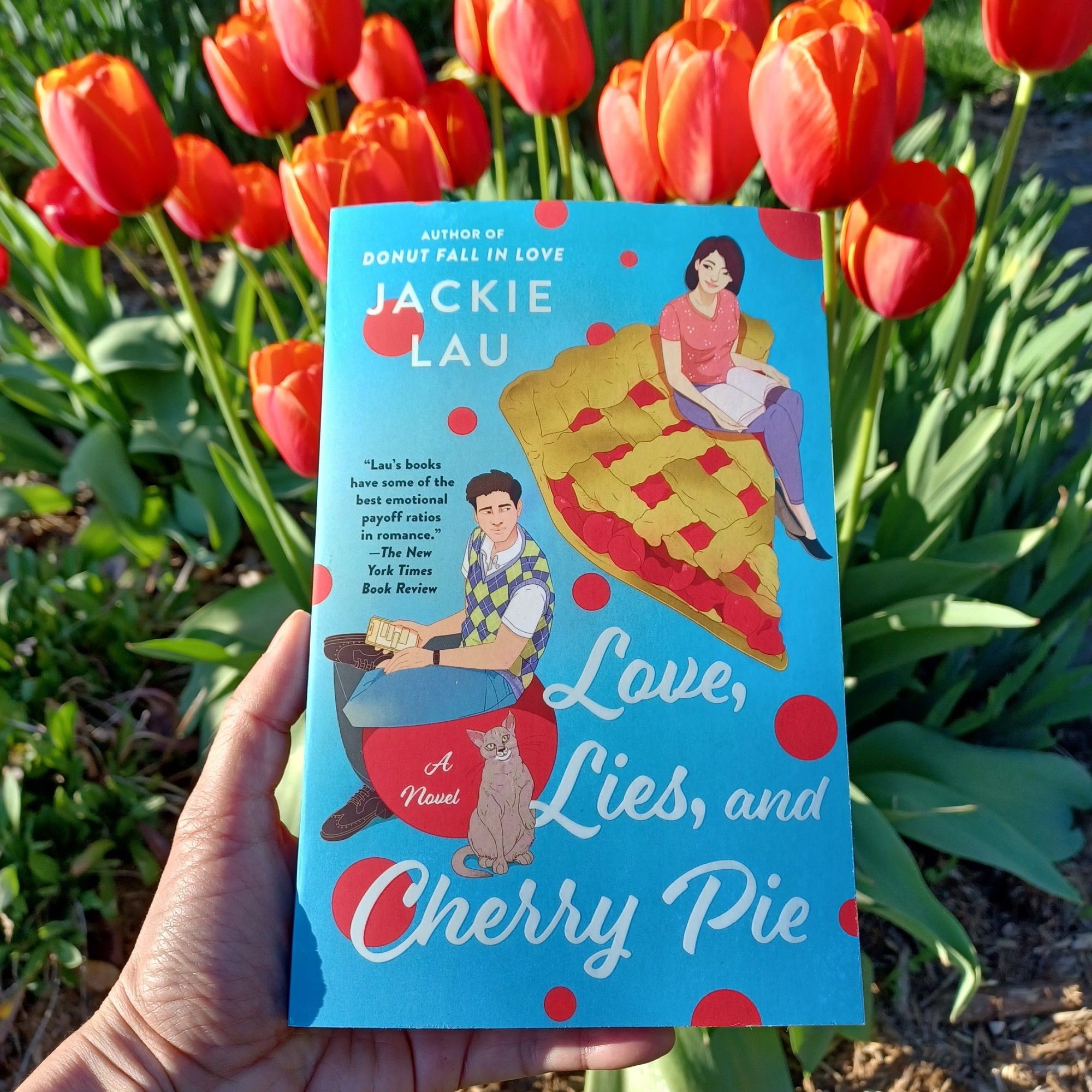 Hand holding paperback of Love, Lies, and Cherry Pie (illustrated cover with blue background) in front of red tulips in a garden.