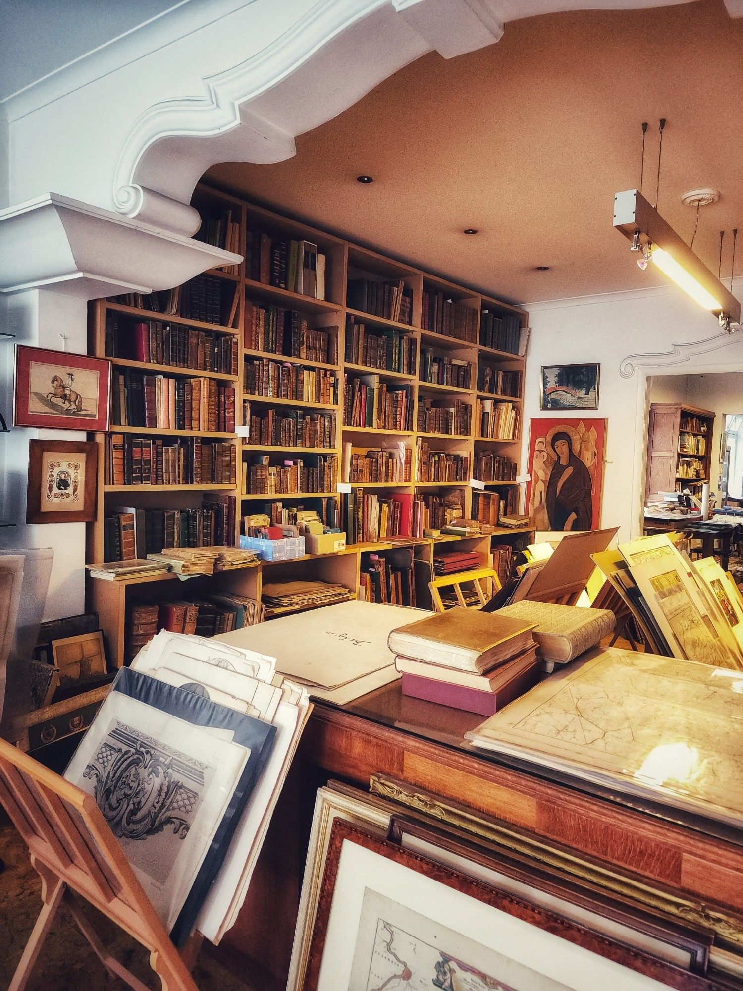 Interior of a shop full of old books, drawings, and prints. In the foreground, stands with large maria and prints and a table with various papers and books. Behind that, a wall covered in bookcases, filled with books of various ages but nothing from the last century or so. Framed art fills the bits of wall that don't have bookcases. A doorway to a room with now books and things in the background.