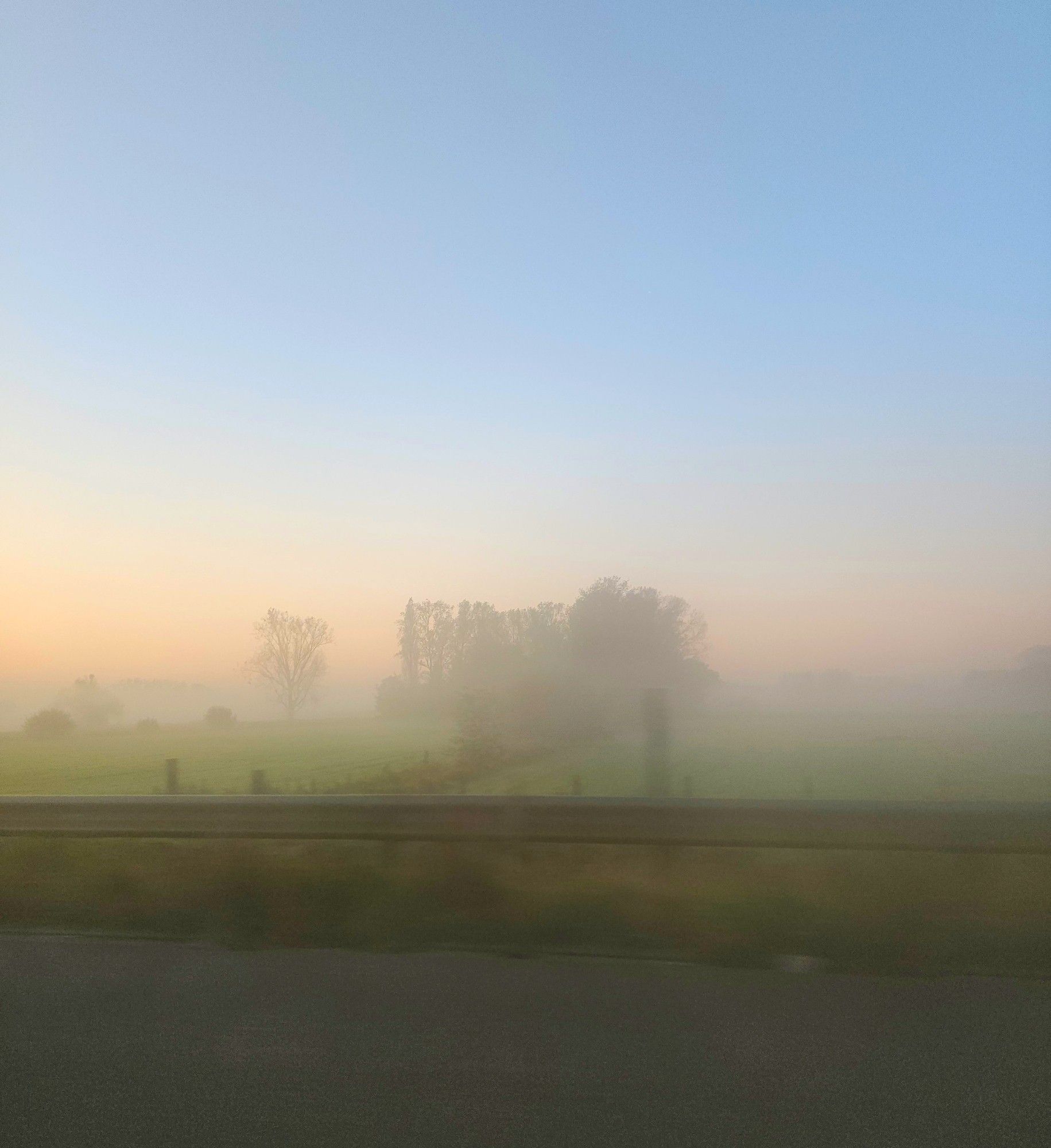 Vertical view through a car passenger window, though the door frame is not included. A very foggy landscape at sunrise. The sky is a soft light blue on top and orange on the horizon, looking all pastel in the fog. Greens fields and trees, looming in the fog are about three quarters down, while the bottom quarter is asphalt and guard rail.