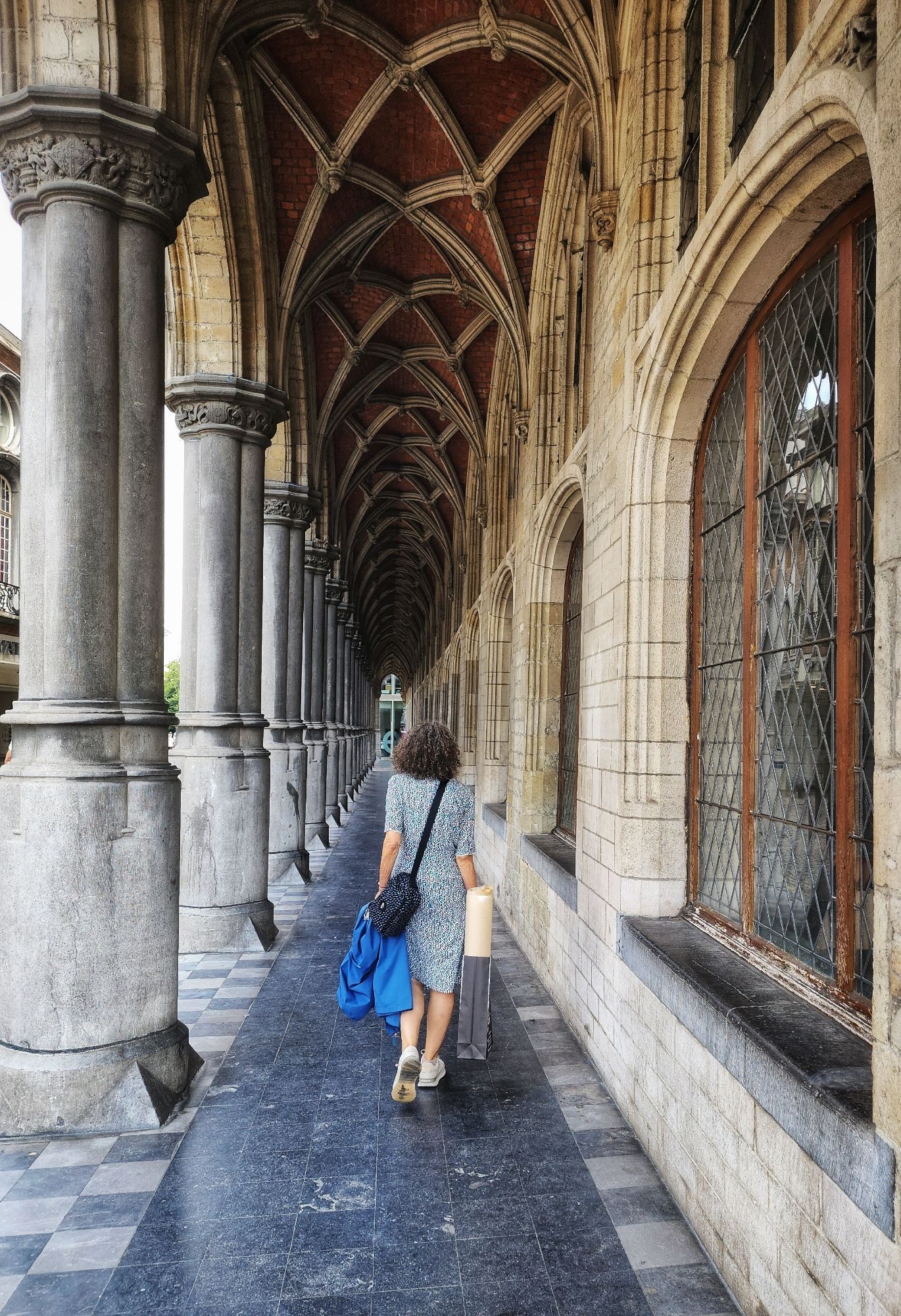 My mom, seen from the back, walking down a covered walkway on the side of the city hall. The floor is shiny dark tile, on the left are light colored pillars, with the street mostly invisible beyond. On the right are windows and above neogothic arches. I believe it's a 20th century construction based on 16th century plans but I'm not positive as the building complex is very complicated. My mom is in a knee length white and blue dress, carrying a small purse, a blue raincoat, and a shopping bag with a roll of paper sticking out.