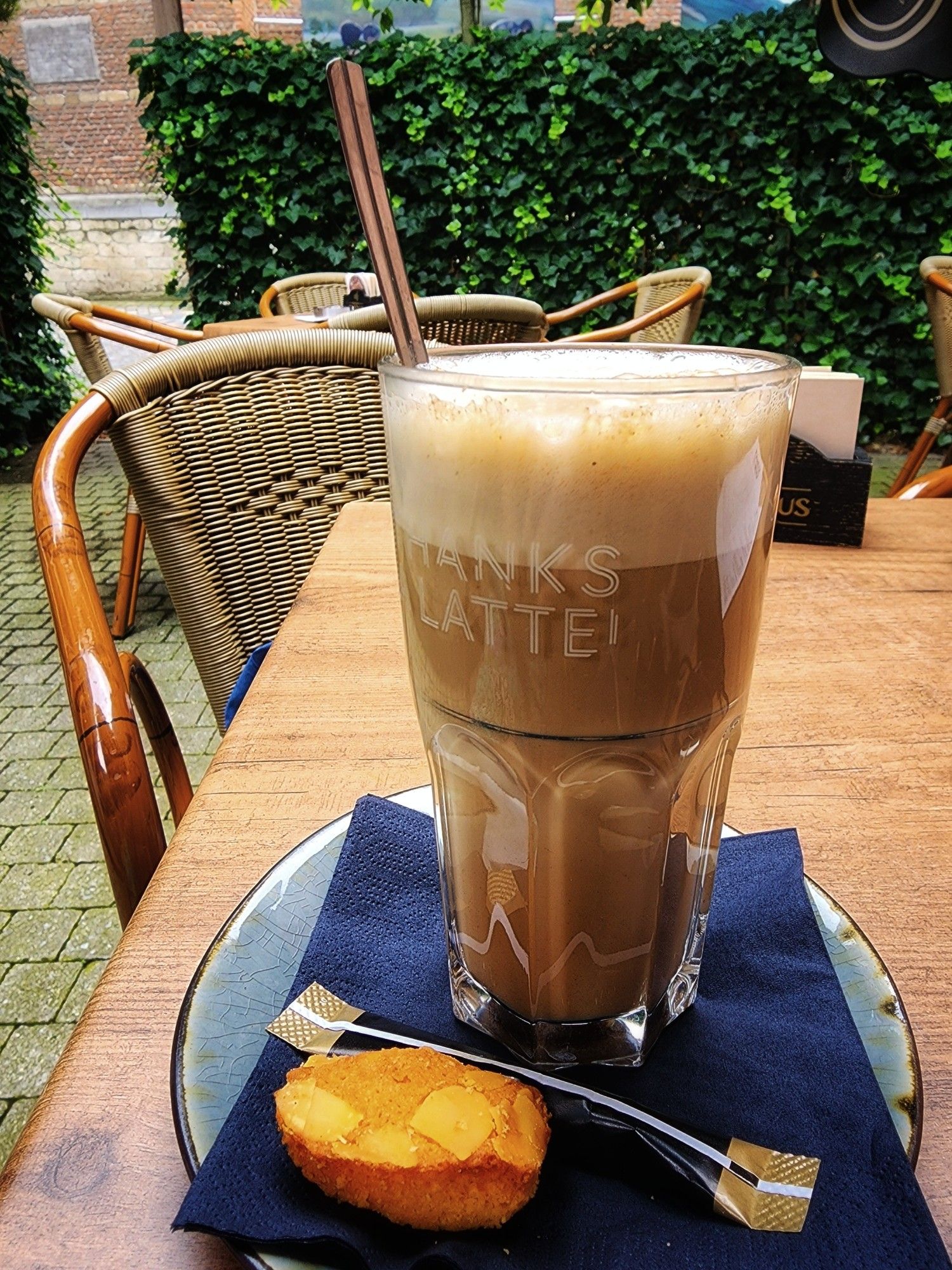 A tall glass of oat milk chai latte, on a small plate with a napkin, sugar, and a small piece of cake. The plate sits on an outdoor cafe table with an empty chair just beyond. Another table sits in the background, beyond that a hedge and gate to a quiet city street with some historic building opposite.