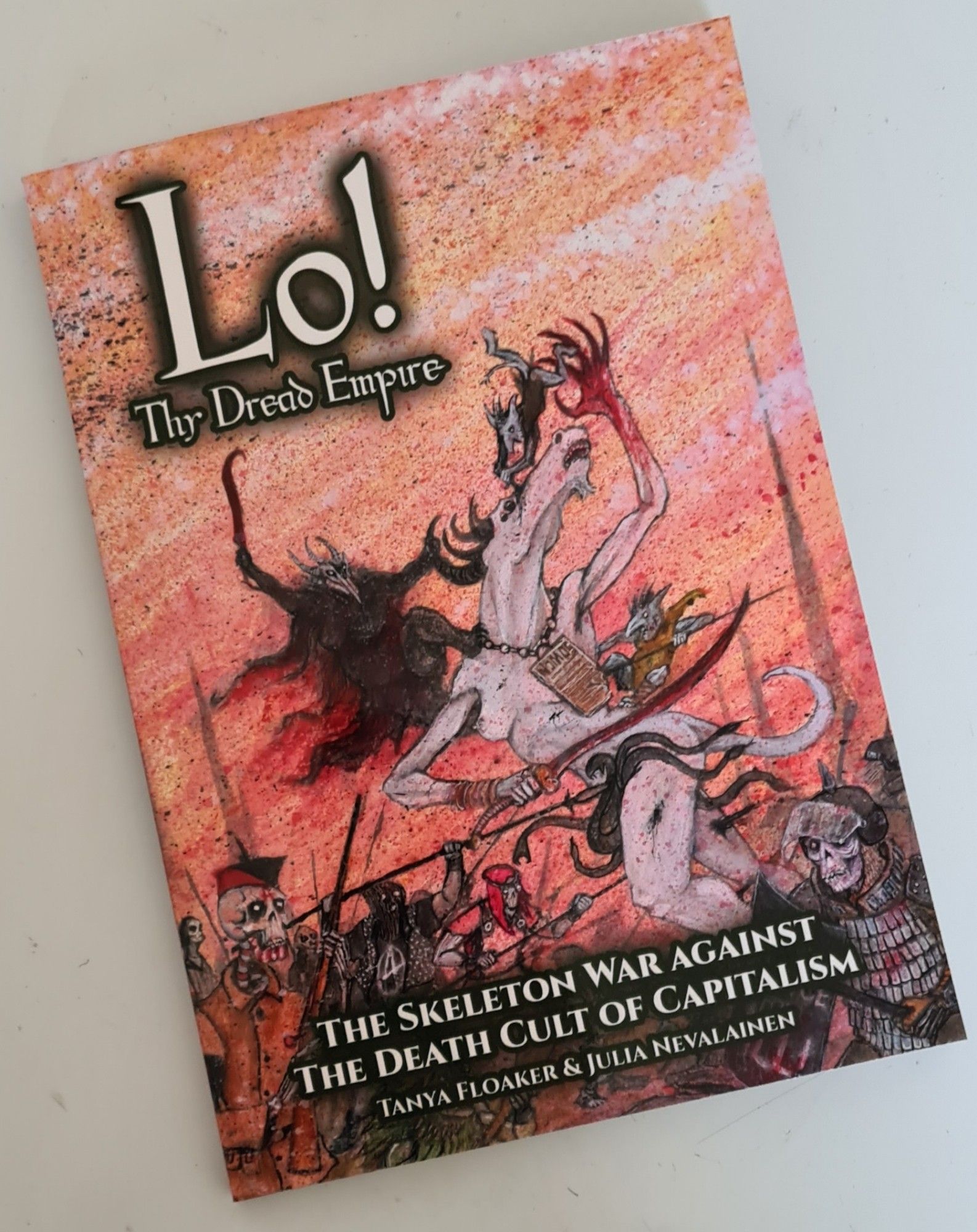 A5 Portrait cover of Lo Thy Dread Empire, with the game name title at the top-left in medieval script font, and the strapline center-bottom of "The Skeleton War against The Death Cult of Capitalism". The art depicts a force of feminist, black bloc and anarcho-syndicalist undead and spirits attacking the demonic foeces of a giant demon of law and riot-police Wights.