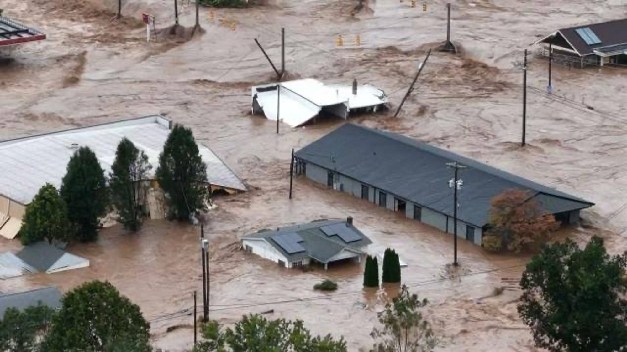 An aerial view of a significant flood, with multiple buildings partially submerged in brown, murky water. Some structures are visibly damaged, and trees and power lines are affected by the floodwaters.