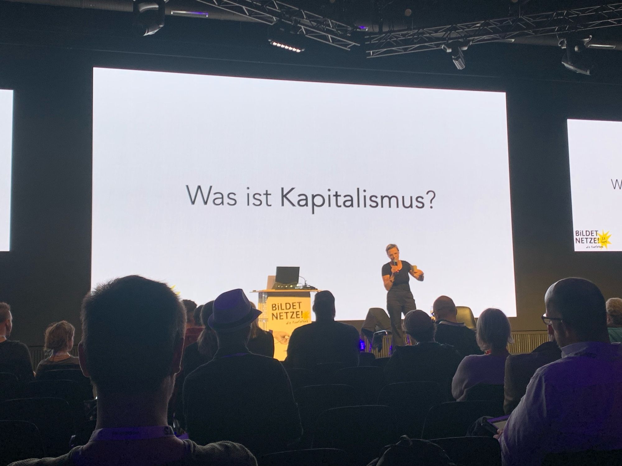 Vortragende, im Hintergrund ein Slide mit der Frage: Was ist Kapitalismus?