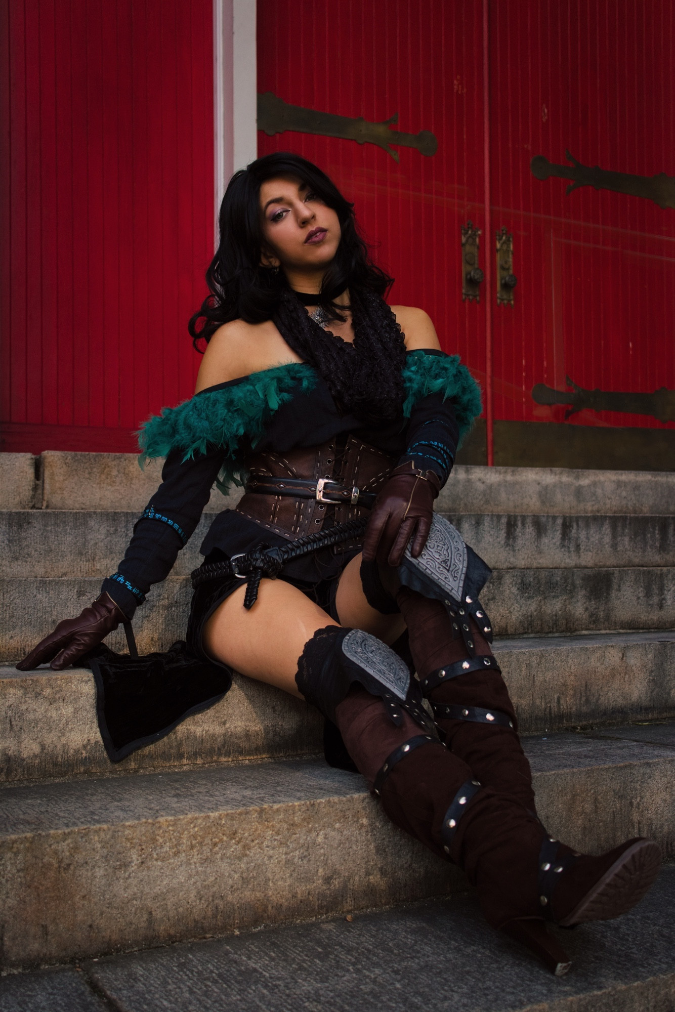 A cosplay of Yennefer (alternative outfit option) from the game, The Witcher 3: The Wild Hunt. The image depicts a person seated on stone steps in front of large red double doors with metal handles. The person is dressed in a fantasy-themed outfit, featuring an off-the-shoulder top adorned with dark green feathers and black fabric. They wear a brown corset with intricate detailing and a wide belt, and their forearms are covered with brown, fingerless gloves. The character’s legs are decorated with brown thigh-high boots featuring silver embellishments and dark fabric accents. Their hair is long and black, cascading over one shoulder. The lighting emphasizes the texture of the clothing and the stone steps.