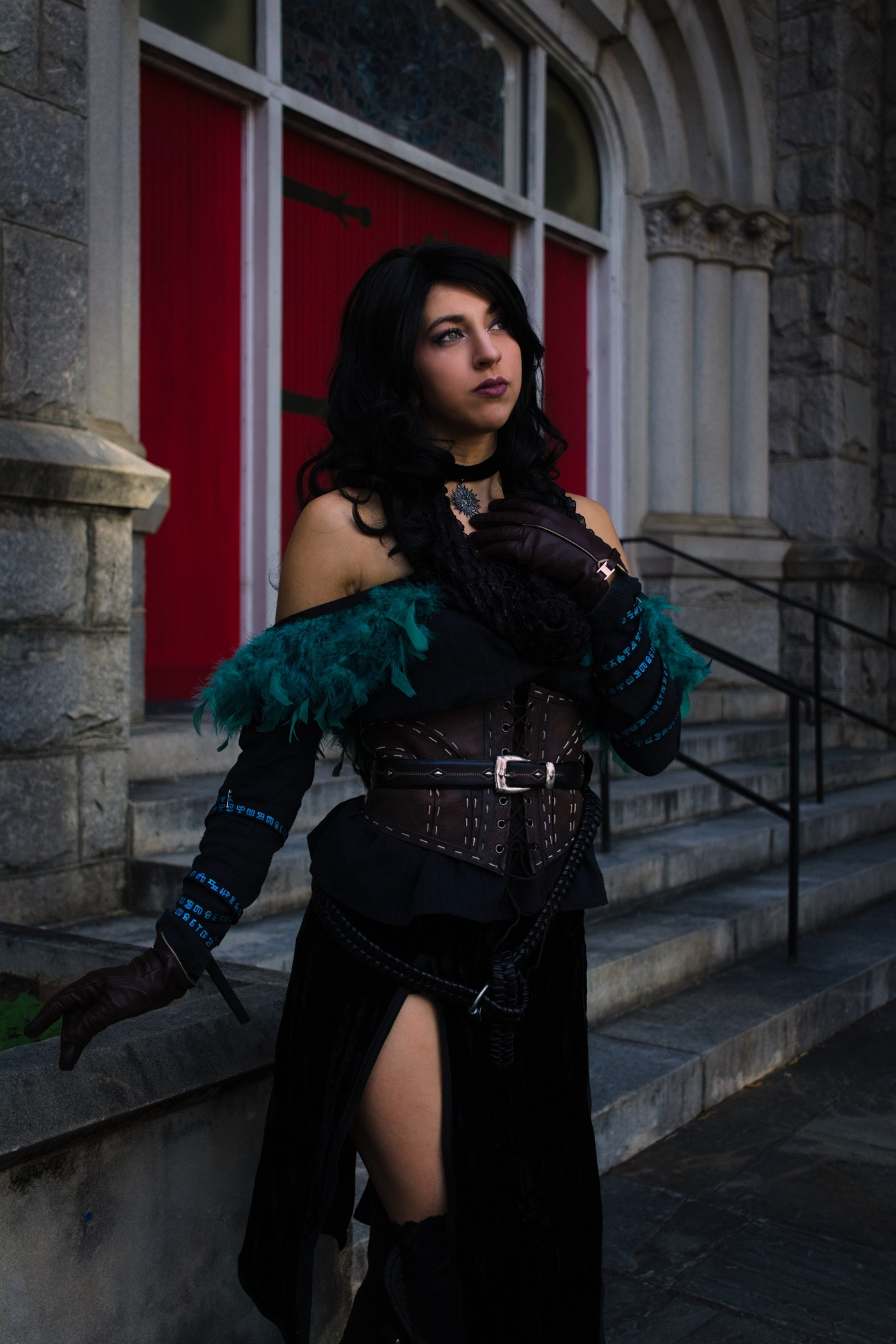 A cosplay of Yennefer (alternative outfit option) from the game, The Witcher 3: The Wild Hunt. A person stands in a dramatic pose outside a stone building, wearing a black off-the-shoulder dress accented with dark teal feathers and a brown leather corset. The corset features several buckles and stitching details. The dress has a high slit, revealing a portion of the left leg. The person wears long, brown leather gloves and holds one hand to their chest, displaying a pendant necklace. Dark hair falls in waves over their shoulders, and their expression is contemplative. Behind them, red doors are set within a gray stone archway. The stonework includes ornate details, and steps with metal railings lead up to the entrance.