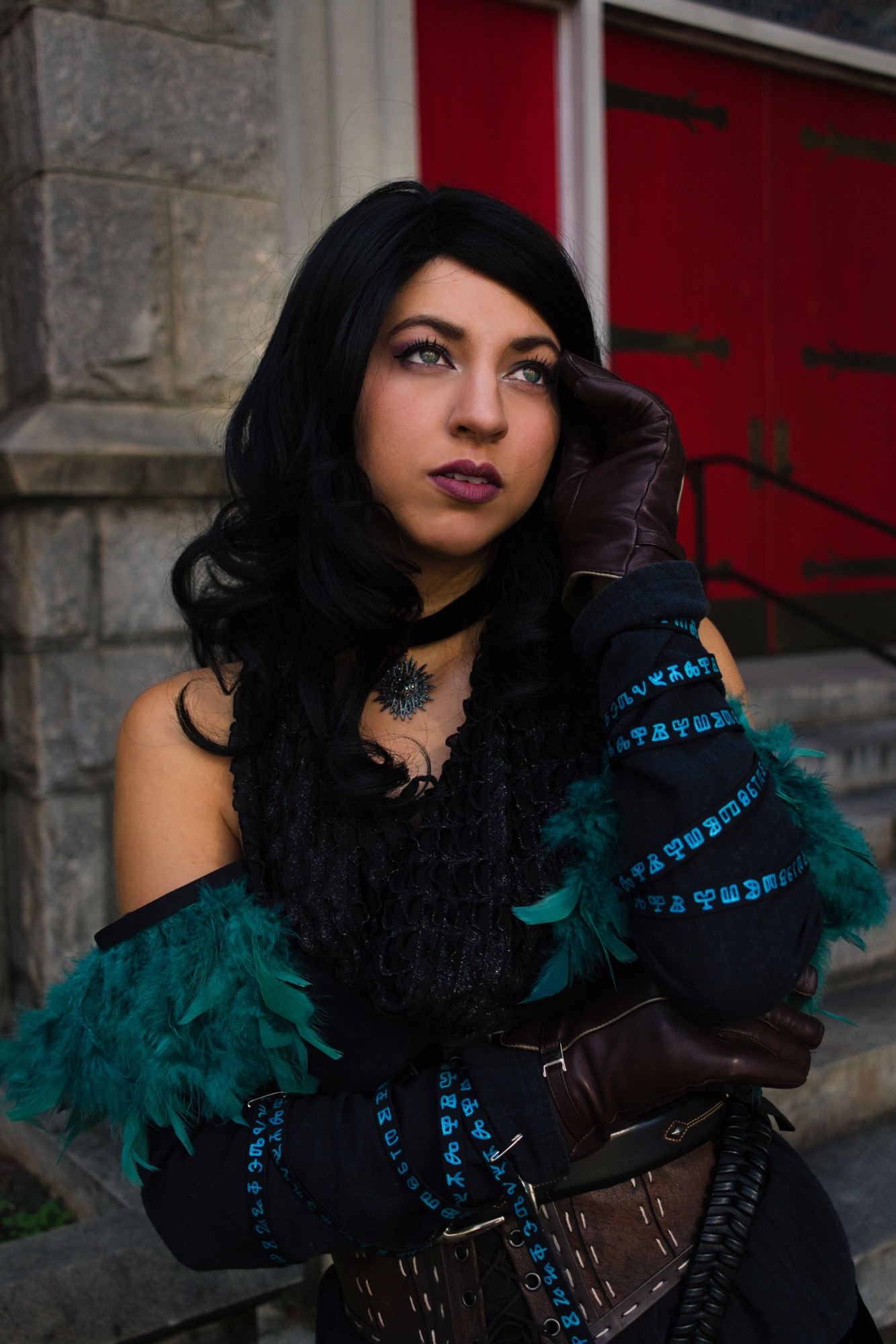 A cosplay of Yennefer (alternative outfit option) from the game, The Witcher 3: The Wild Hunt. The image portrays a person standing outdoors, dressed in a detailed costume. The individual has long black hair and is wearing dark makeup, including deep maroon lipstick and eye makeup that highlights their eyes. They are wearing a black textured top with a feathered teal trim along the sleeves. Their gloved hand is positioned near their face in a contemplative pose. The gloves are brown leather, complemented by an arm covering with bright blue symbols or characters. A pendant with a spiky design is visible around their neck. The background features a stone building with distinct red doors accentuated by black ironwork details.