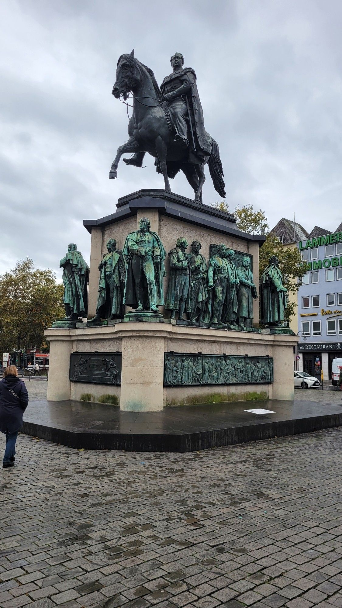 A large statue in the courtyard in Germany. There's a large statue of a man on a horse. Under this statue are several copper statues of various men