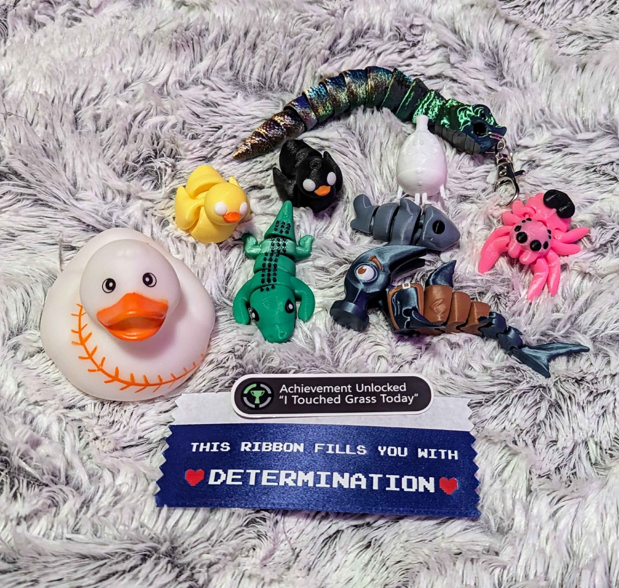 Baseball rubber ducky, Yellow 3D printed duck fidget toy, Black 3D printed duck fidget toy, Green alligator fidget toy, Duo chrome hog nose fidget keychain, White Egg Boy, Gray shark fidget toy, Pink 3D printed spider with black eyes and heart fidget toy, Gray and brown hammer head shark (head shaped like the tool hammer) fidget toy, Achievement Unlocked "I Touched Grass Today" sticker, This Ribbon Fills You with Determination blue ribbon