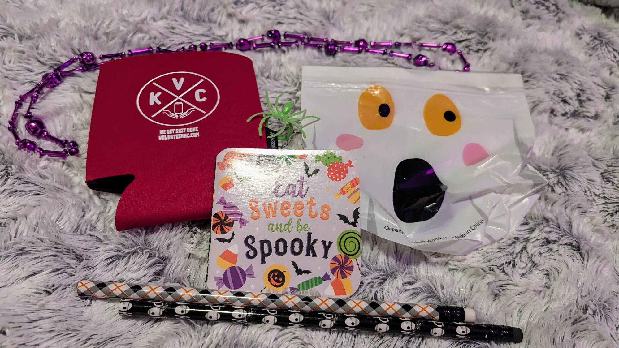 Red can cooler sleeve with KVC logo, Spooky themed sweets tiny booklet, Fluorescent green spider ring, White ghost bag with yellow eyes black pupils pink blush and black 0 mouth, Two patterned wooden pencils