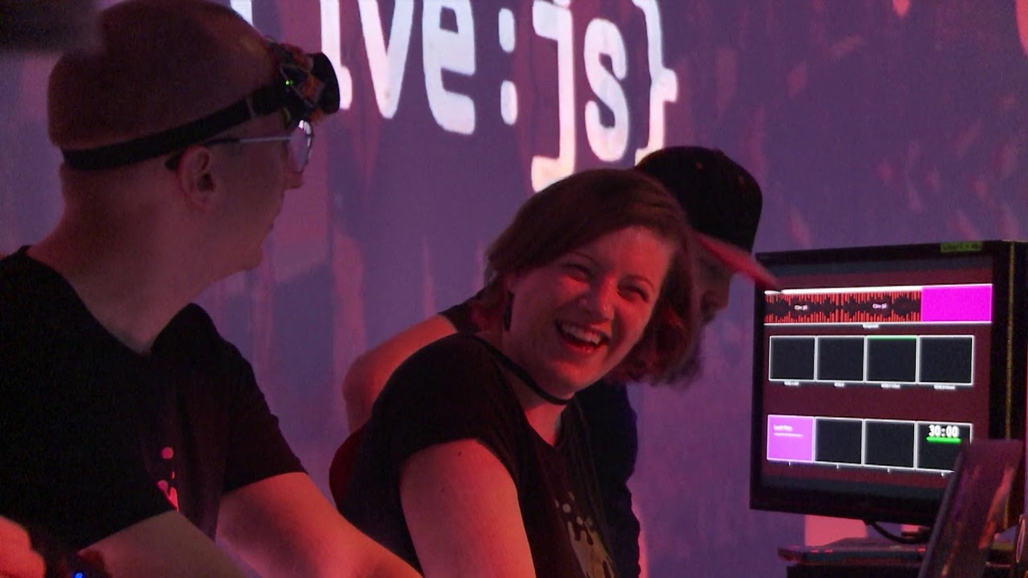 Tim Pietrusky, Ruth John and Martin Schuhfuss are standing on stage at JSConf EU, doing the intro performance while smiling at each other. In the background is a small piece of the projection surface of the sage, showing the LiveJS logo. 