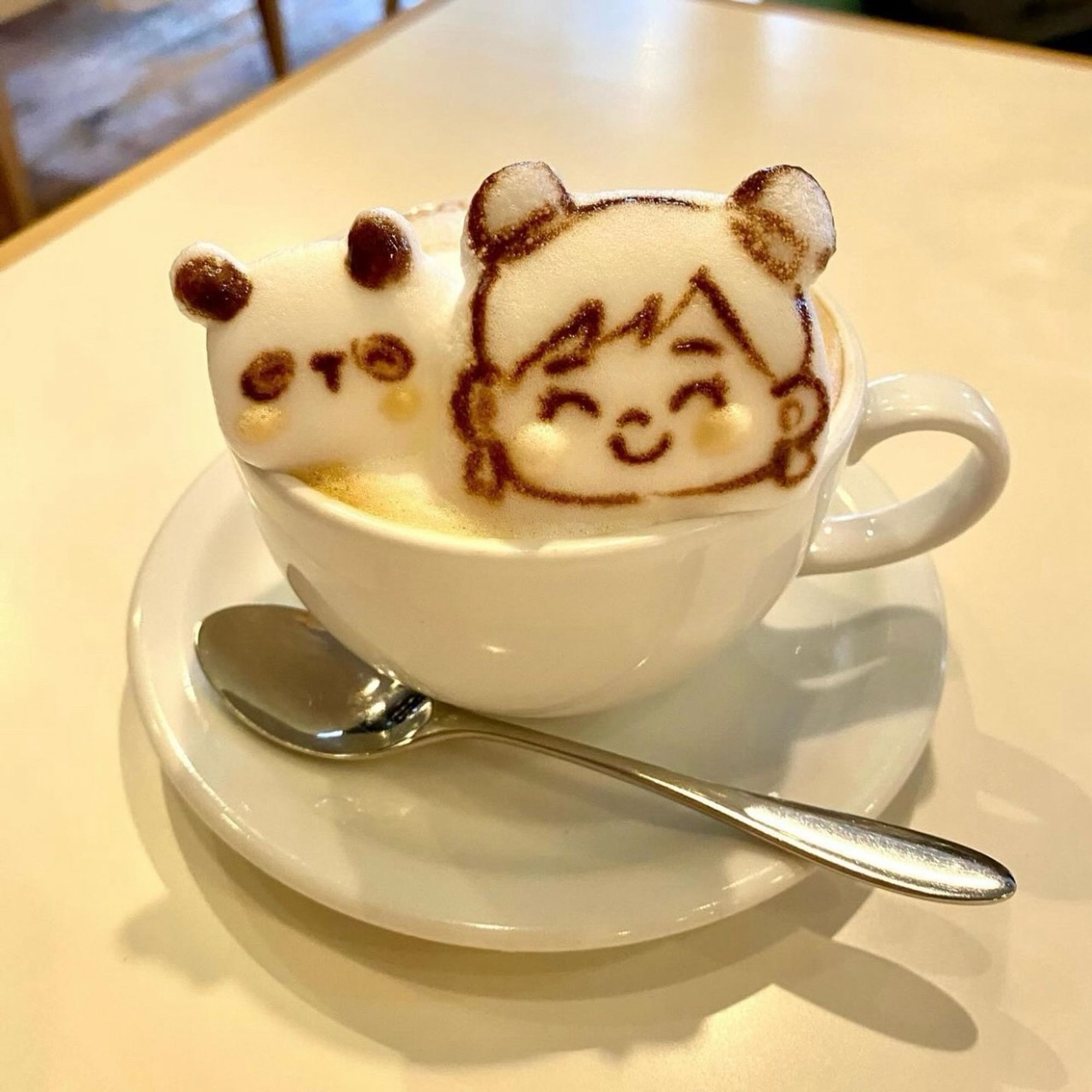 A cafe latte 3d foam art of Lyn Lyn (my book’s main character) and her panda plushie friend inside a mug with a spoon on 
the side