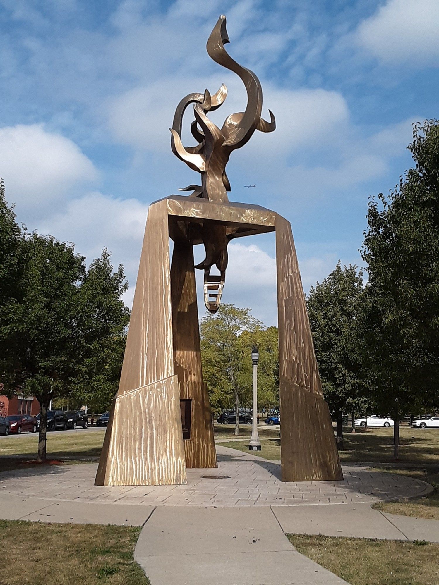 A large, abstract bronze sculpture stands in an outdoor park setting. The sculpture is composed of three tall, textured, spire-like pillars supporting an intricate, swirling design at the top. Trees, a lamppost, and a pathway are visible in the background under a partly cloudy sky. This is a monument dedicated to Ida B Wells in Chicago.