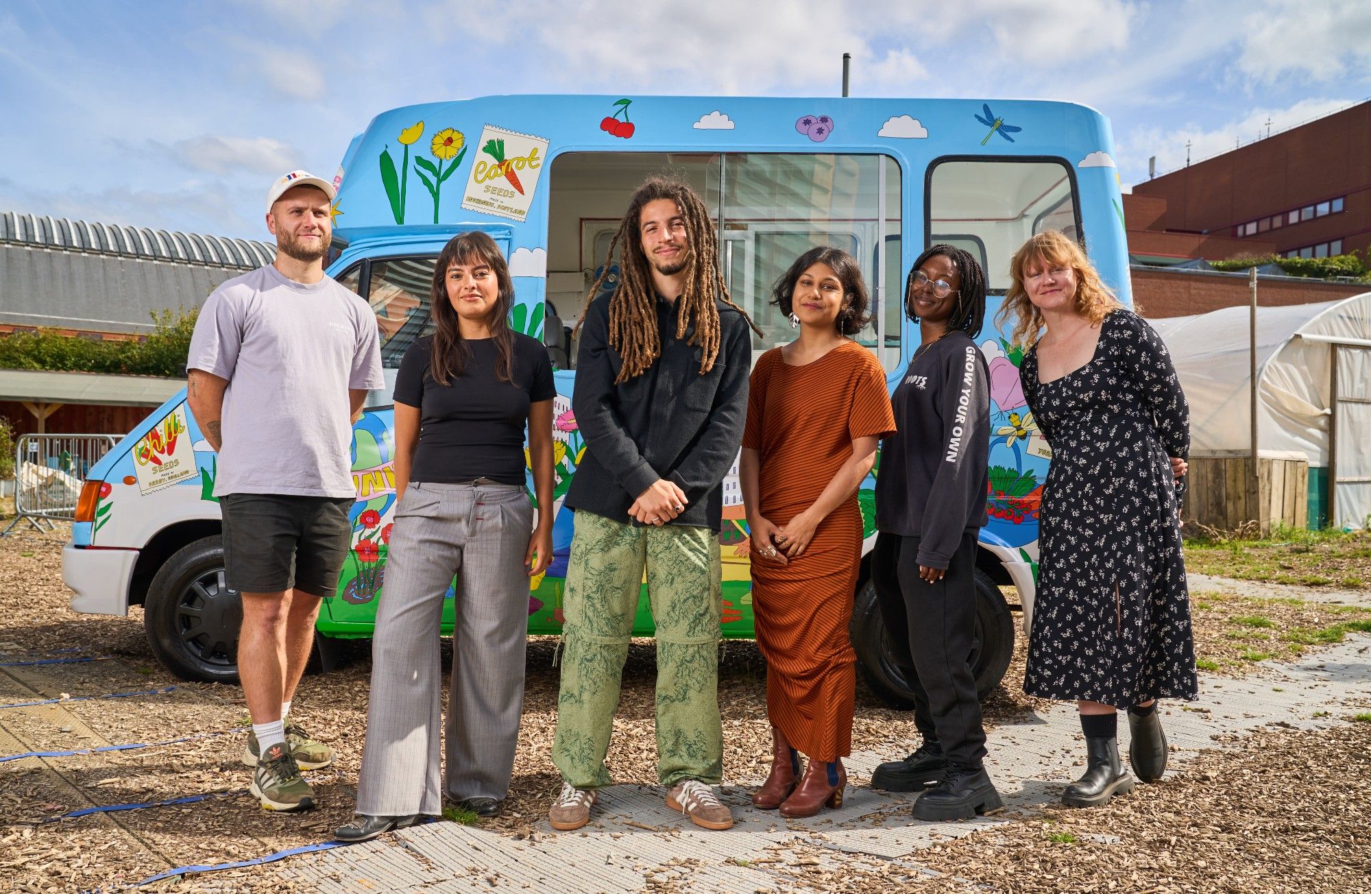 Six people are stood next to each other smiling at the camera. Behind them is a van - it looks like an ice cream van - with images of fruit, clouds, flowers and bugs over it.