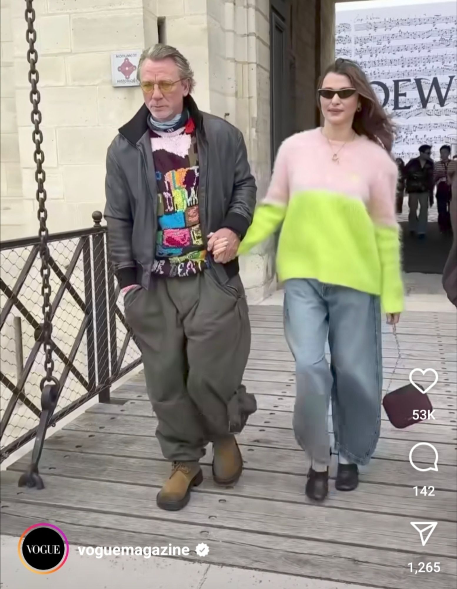 daniel craig and rachel weisz at the loewe show, screengrab of a video by vogue. daniel is dressed like a teen dj in 1987 and rachel is dressed in light baggy jeans and an oversized pink and green colorblocked sweater