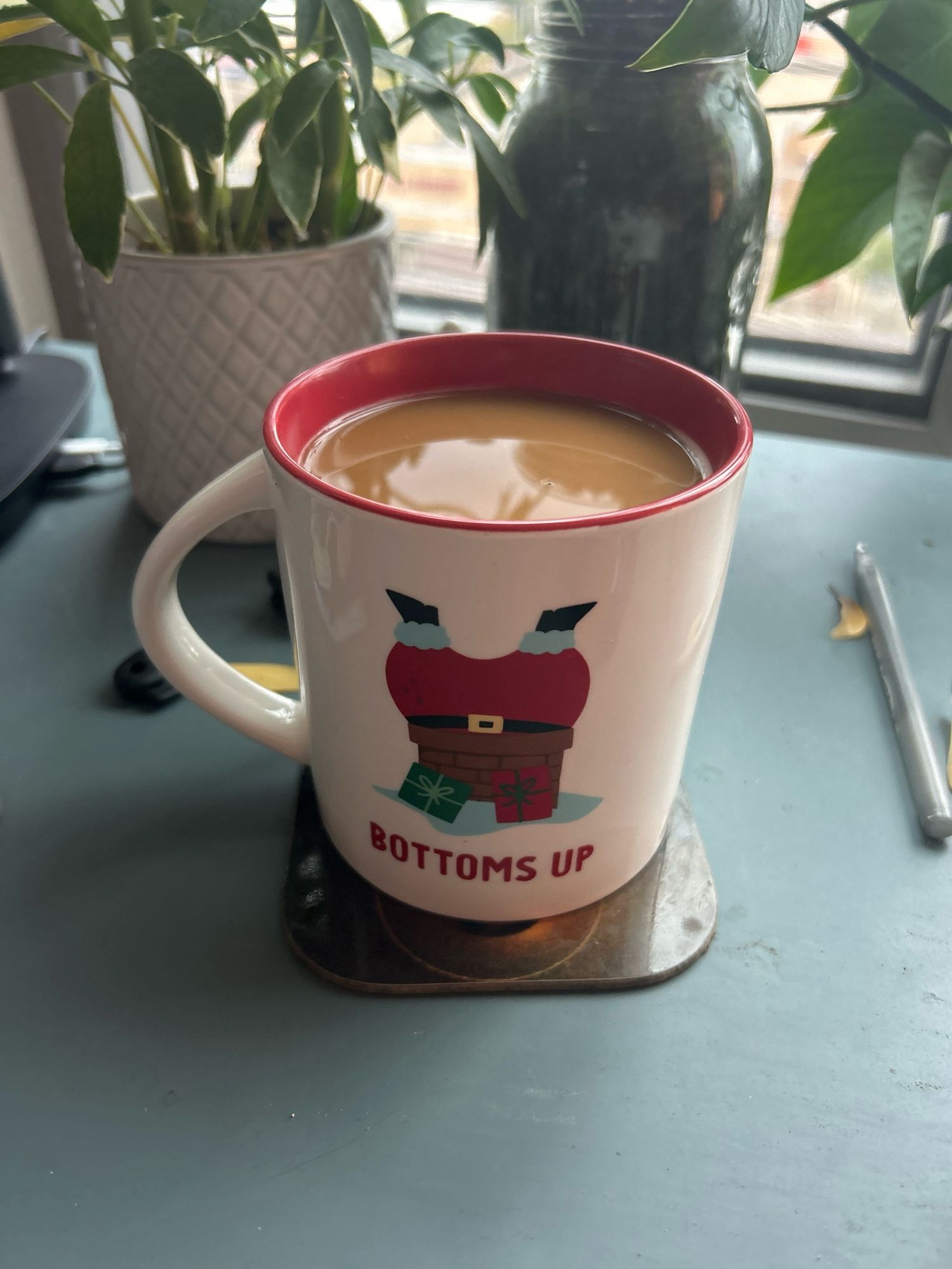 This users coffee in a white mug with Santa’s ass and gams hanging out of a chimney with the phrase “bottoms up” below sitting on a coaster in front of a schefflera and a pothos