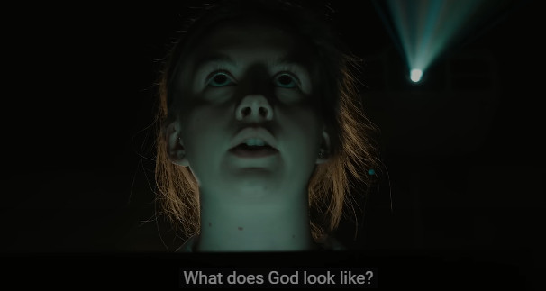 Num ambiente escuro, a protagonista do vídeo olha para cima, com o rosto iluminado por baixo, e pergunta em inglês, a um elocutor oculto, "what does God look like?", texto que aparece na legenda do vídeo pouco abaixo.