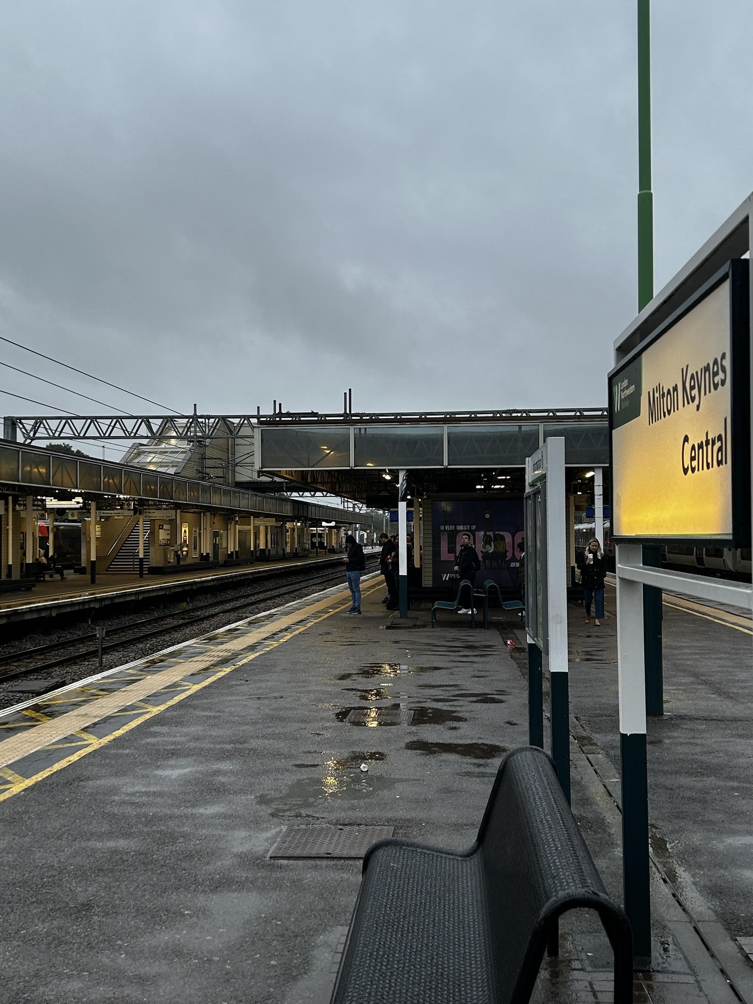 Picture of the illuminated station sign in a platform at Milton Keynes in grey, wet weather 