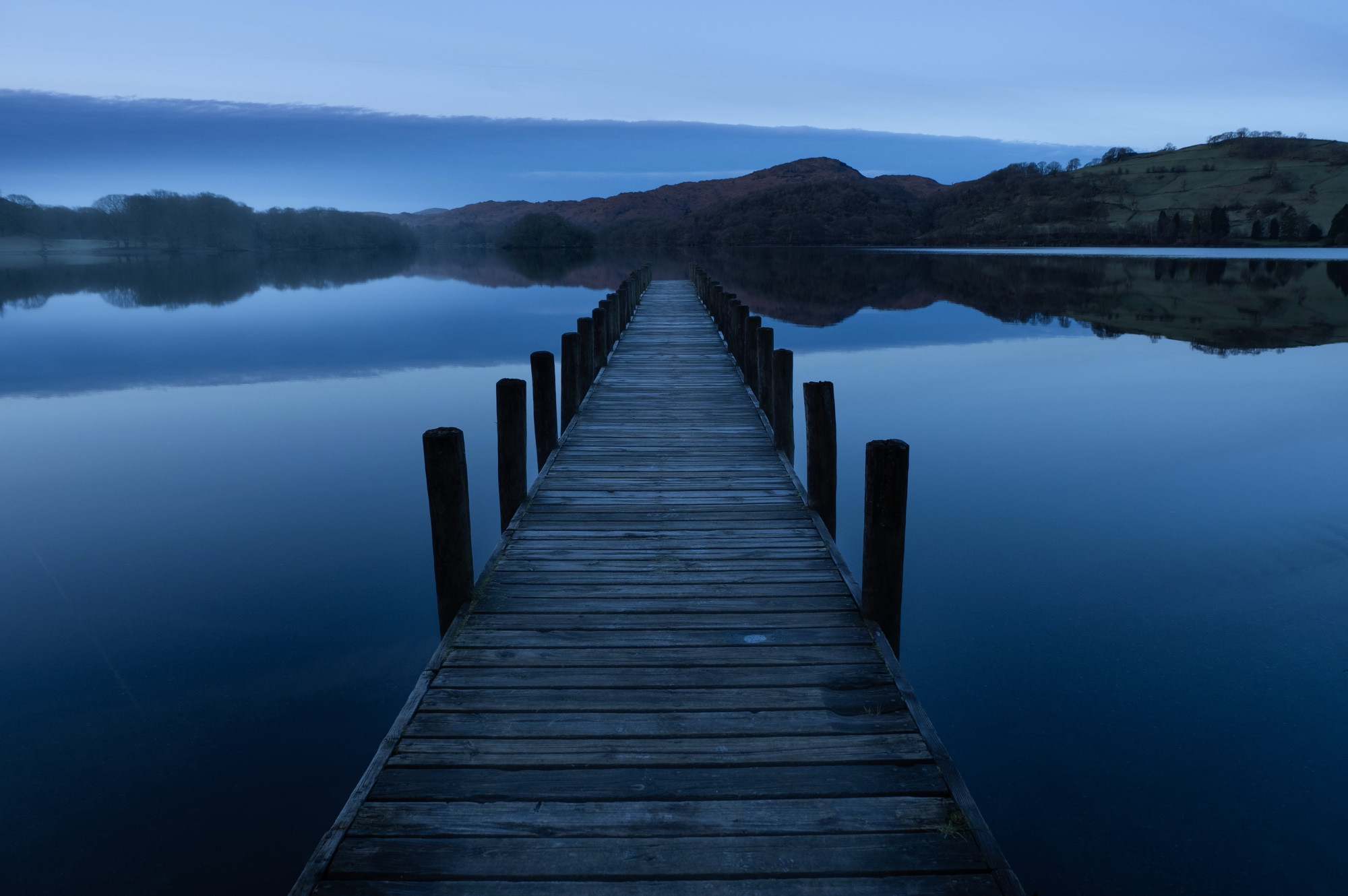 A wooden jetty runs out into a calm lake, it appears to run almost to the horizon. 