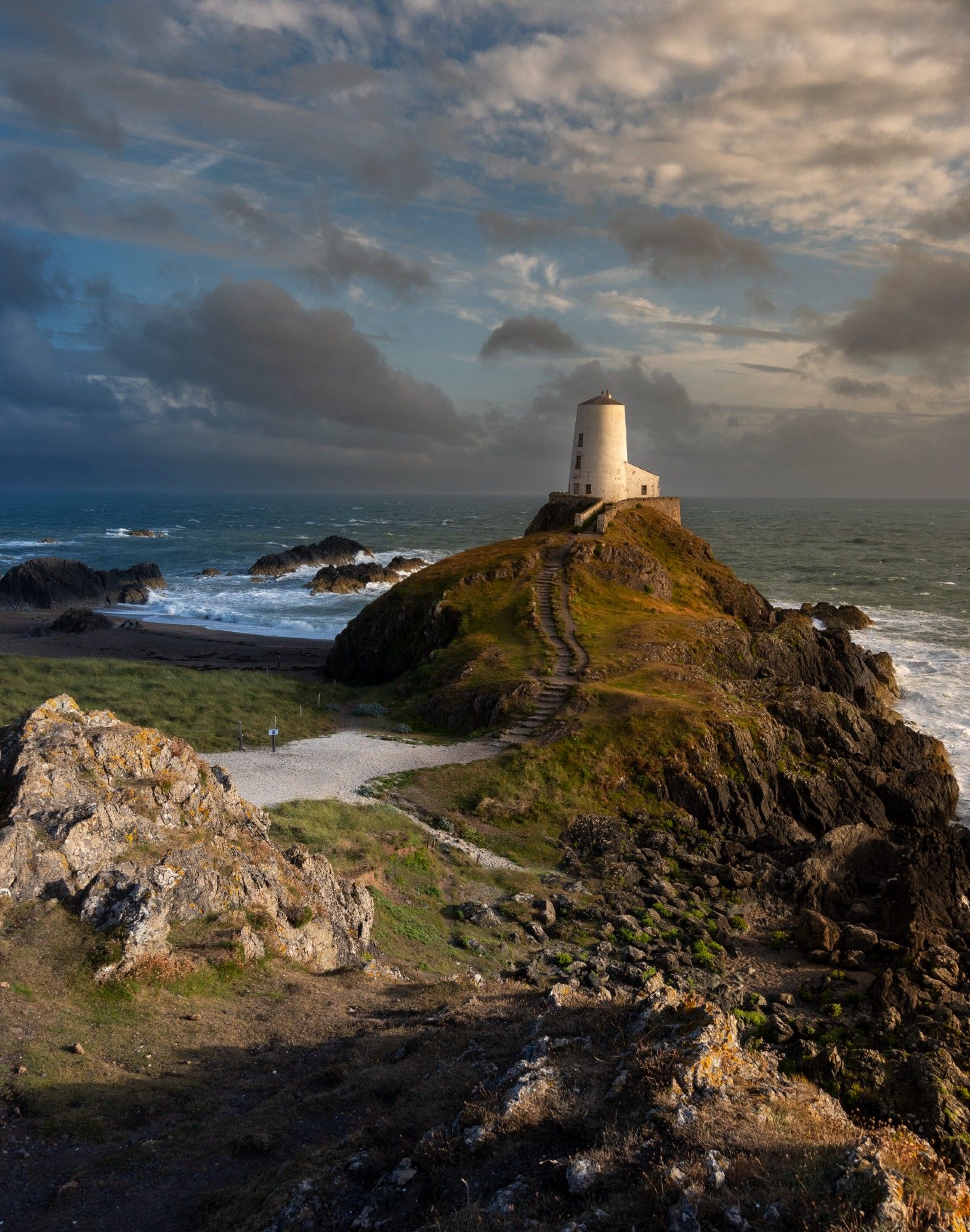 A white painted lighthouse stands on top of a rocky outcrop with stairs leading up to it. Behind the sea is rough with white waves crashing against the shore. The sky is partly cloudy with a golden glow from the sun setting to the right of the frame.