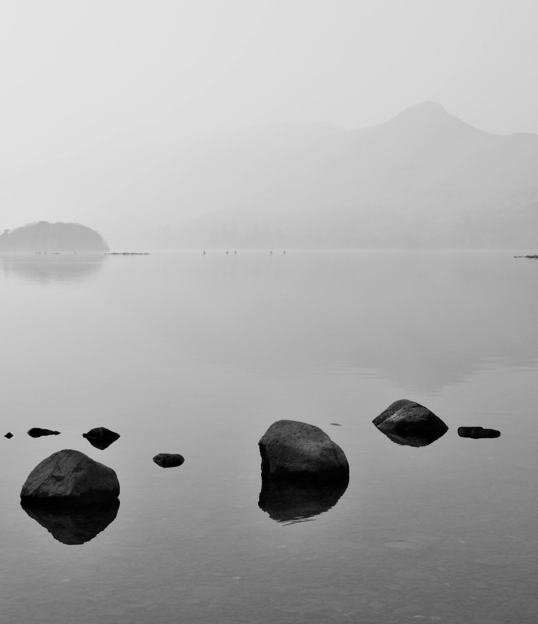 Rocks stand in shallow calm lake waters in the foreground. in the background an island and canoes can be seen through the mist that obscures the far side of the lake. Above the lake the distinctive peak of Catbells is just visible.