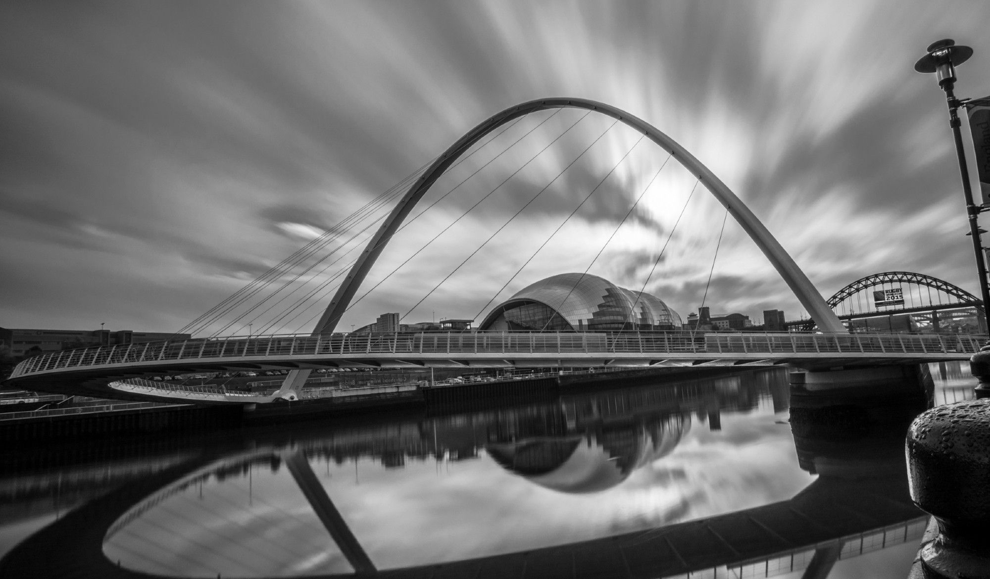 A black and white photo of a metal arch bridge across a calm river. The sky above the bridge is streaked by clouds moving on a windy, their movement caprured by the use of a long exposure.