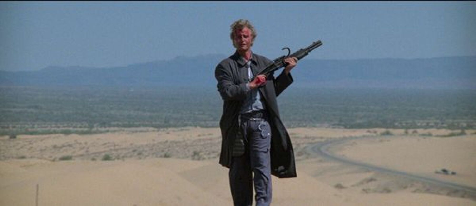 A bloodstained, scruffy but confident-looking white man with a shotgun walks out of a desert.